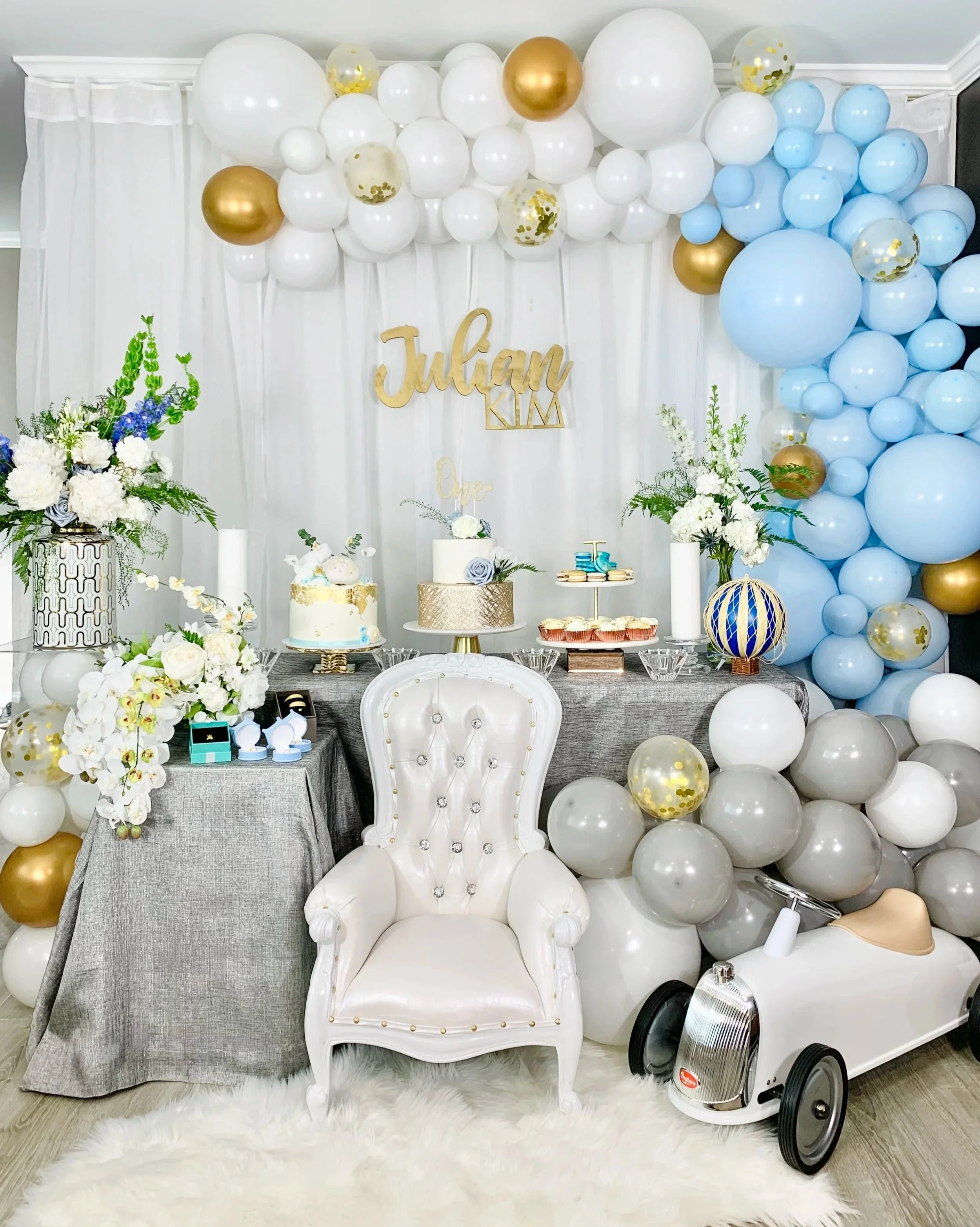 A decorated party setup with a white and gold balloon arch, white floral arrangements, and a white high-back chair in the foreground. The backdrop has a gold sign that reads "Julyan" with smaller signs underneath. There are decorated cakes, cupcakes, and a balloon garland on the table. The setting has a white fur rug and a white curtain backdrop.