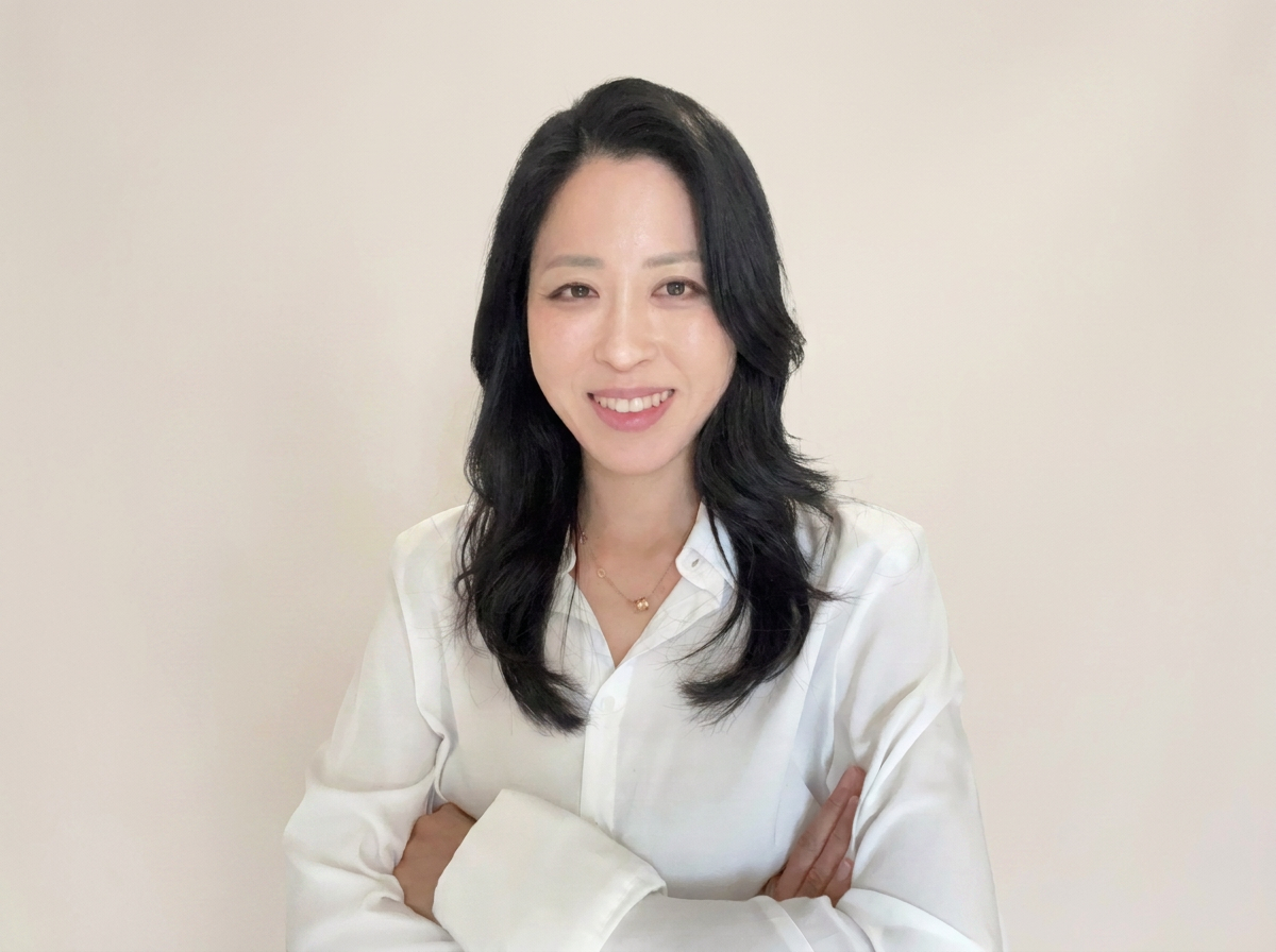 A woman with long black hair, wearing a white button-up shirt, smiling with her arms crossed, standing against a plain light-colored background.