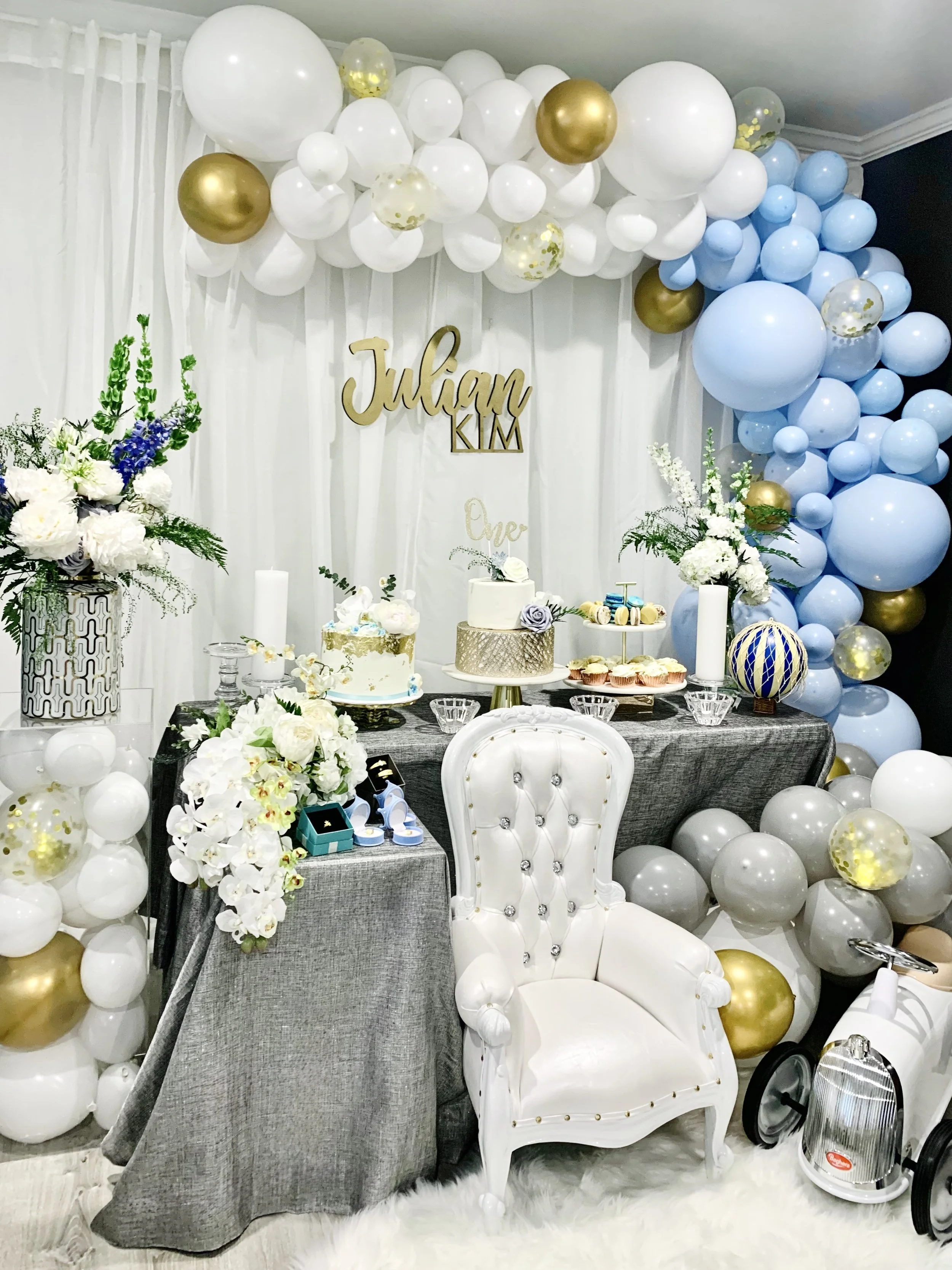A decorated celebration area with white, gold, and blue balloons forming an arch and backdrop, a white floral arrangement, a cake on a stand, and a white ornate chair in front. There are small gift boxes, cupcakes, and a vintage-style fan on the tabl