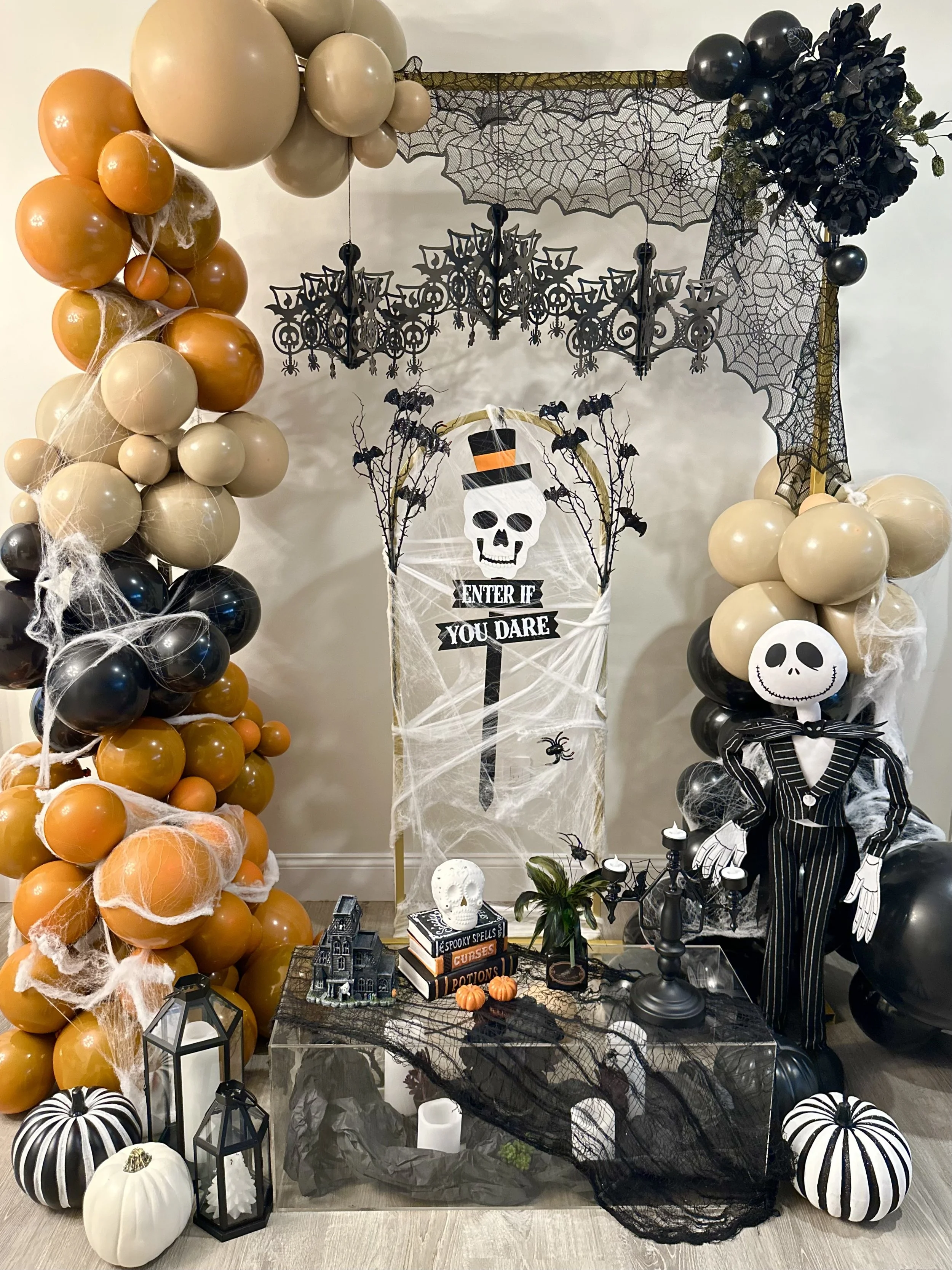 Halloween themed decoration with black, white, orange, and beige balloons, cobwebs, black lanterns, skulls, miniature pumpkins, books, a framed skeleton, and a table covered with black lace and spider decorations.