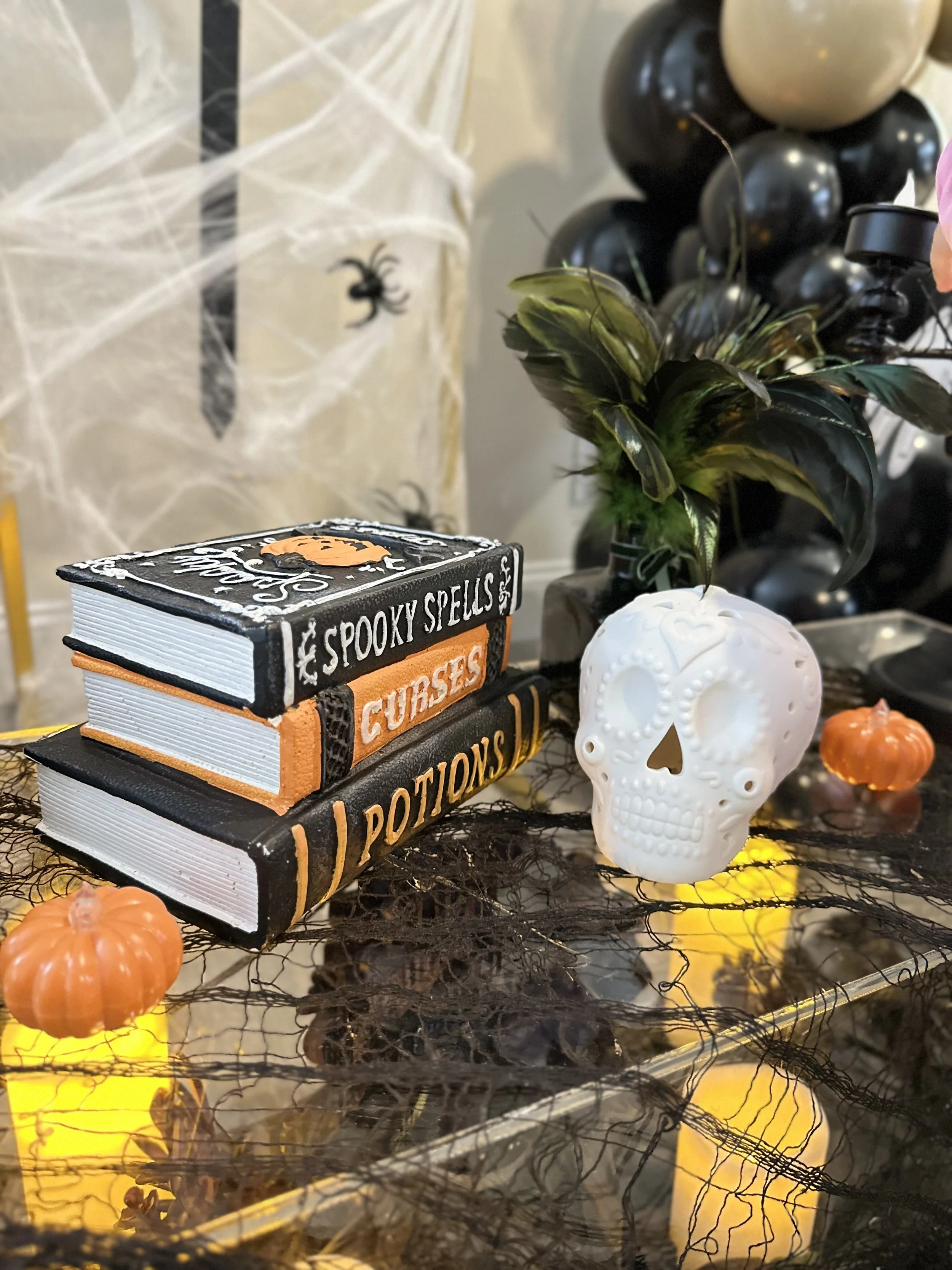Halloween decorations including a white skull, a stack of books labeled 'Spooky Spells' and 'Potions', a black and orange spider decoration, a large plant with dark leaves, a cluster of black and beige balloons, small orange pumpkins, a spider web, a