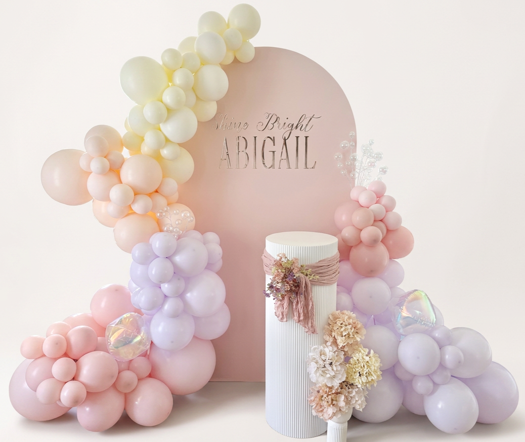 Decorative backdrop with pastel balloons, floral arrangement, and a pink sign that reads 'Shine Bright Abigail' for celebration.