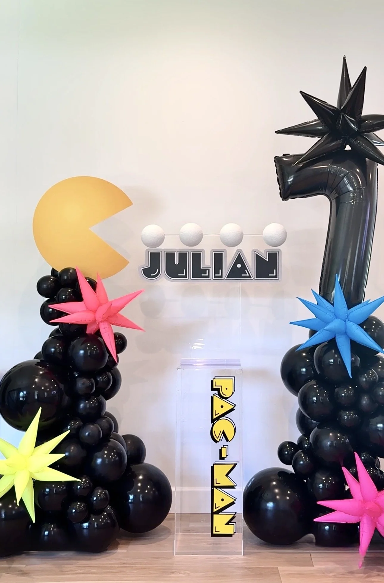 Decorative balloon setup with black balloons forming columns, colorful star-shaped balloons, a Pac-Man themed yellow balloon, a black number balloon, a black starburst balloon, and a sign with the name 'JULIÁN' in black, white, and gray.