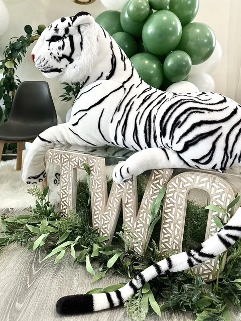 A white tiger with black stripes lying on a decorative table surrounded by green plants and balloons, with black chairs in the background.