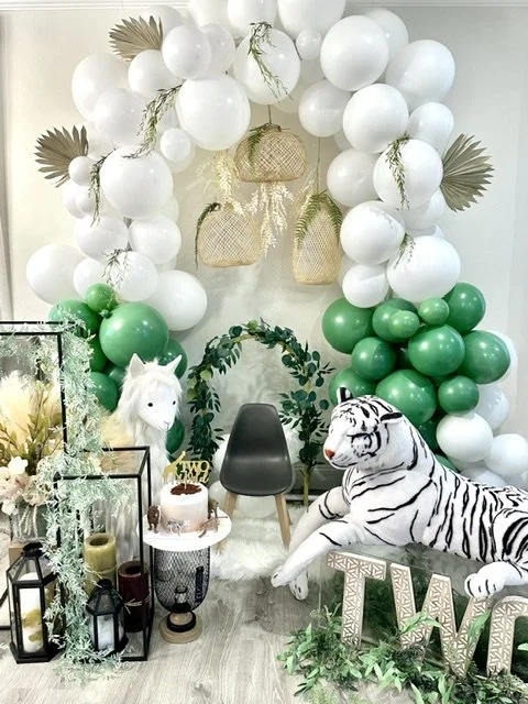 Decorative setup with white and green balloons, a white plush unicorn, a black chair, a large white plush tiger, greenery, lanterns, and floral arrangements, suggesting a celebration or event space.