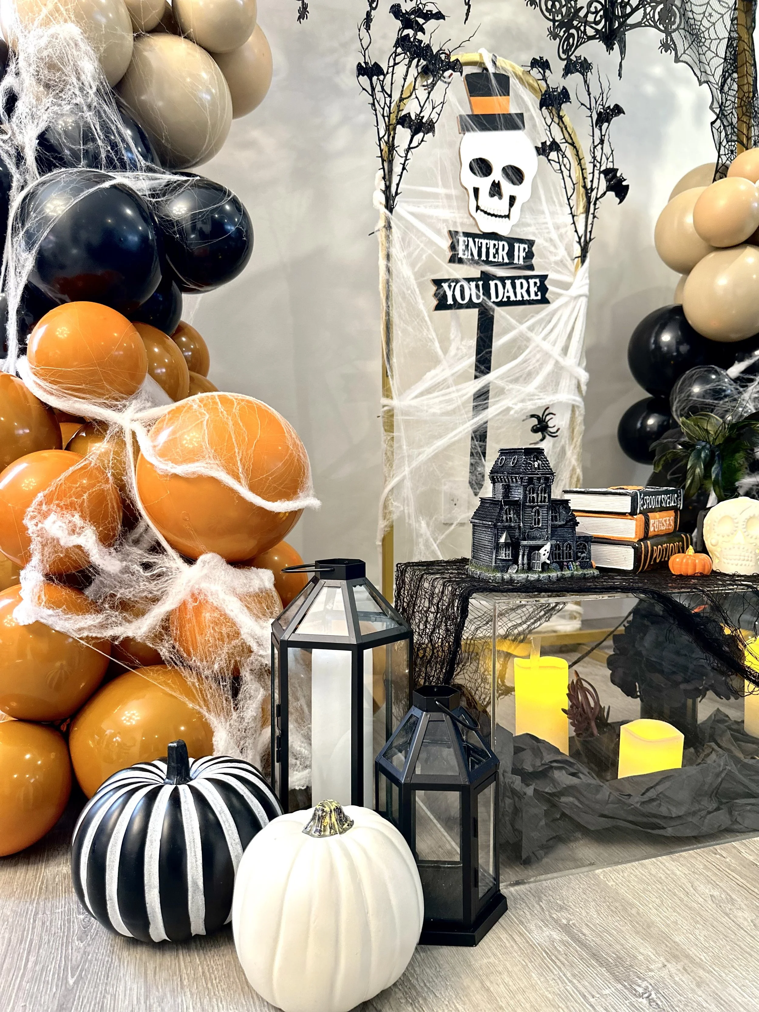 Halloween decorations include black, orange, white, and silver pumpkins, black and orange balloons with cobwebs, black lanterns, a haunted house figurine, candles, spider webs, and spooky sign with a skull and bats.
