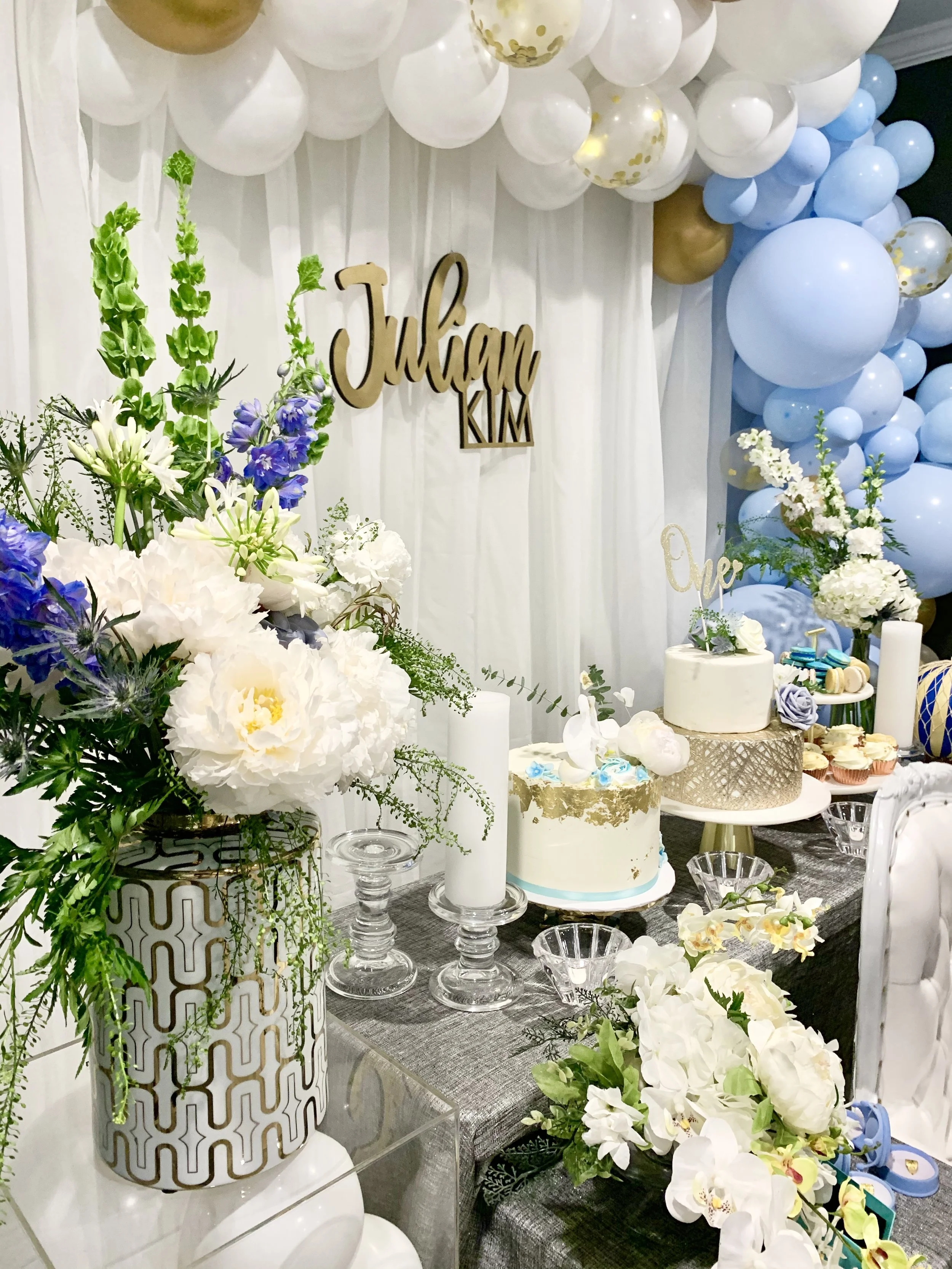 A decorated celebration table with balloons, flowers, and cakes. There is a wooden sign with the names 'Julian Kim' on a white backdrop, and various white, blue, and gold balloons arranged above and around the table.