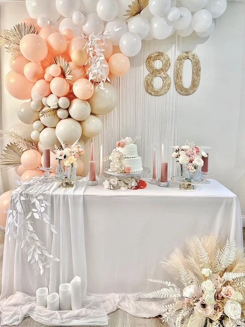 Decorated birthday celebration table with balloon arch, large gold '80' numbers, cake, floral arrangements, and candles.