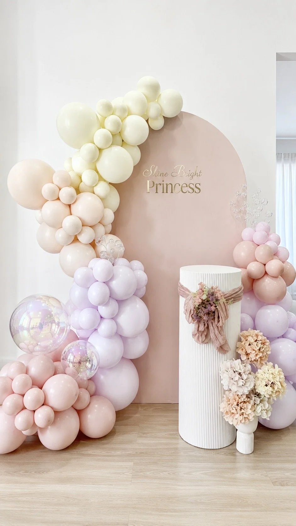 Decorative setup with pastel-colored balloons, a pink backdrop with the words "Shine Bright Princess," and a white cylindrical pedestal adorned with pink fabric and floral arrangements for a celebration or event.