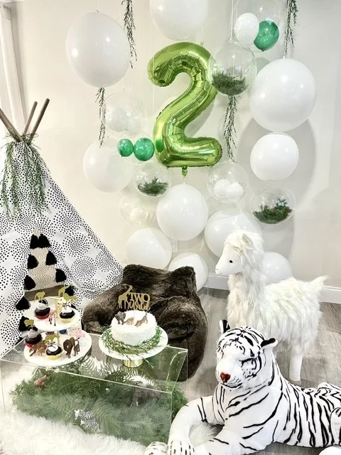 Birthday party setup with white and green balloons, a large metallic green number '2' balloon, a white plush unicorn, a zebra stuffed animal, a decorated cake, cupcakes, and decorative trees.