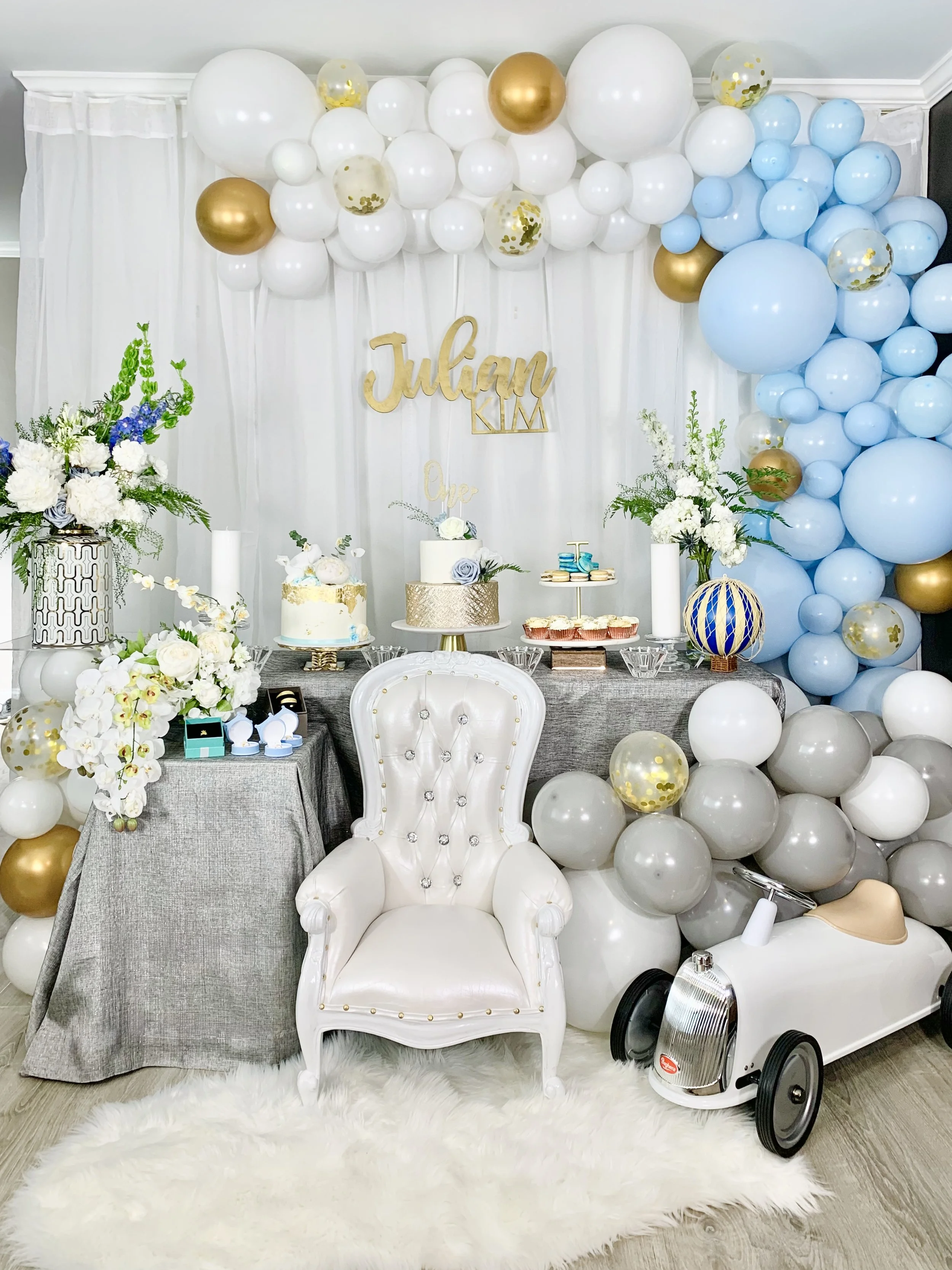 Party setup with white, gold, blue balloons, floral arrangements, birthday cake, and a white ornate chair, with a backdrop of white curtains and a banner with the name Julian.