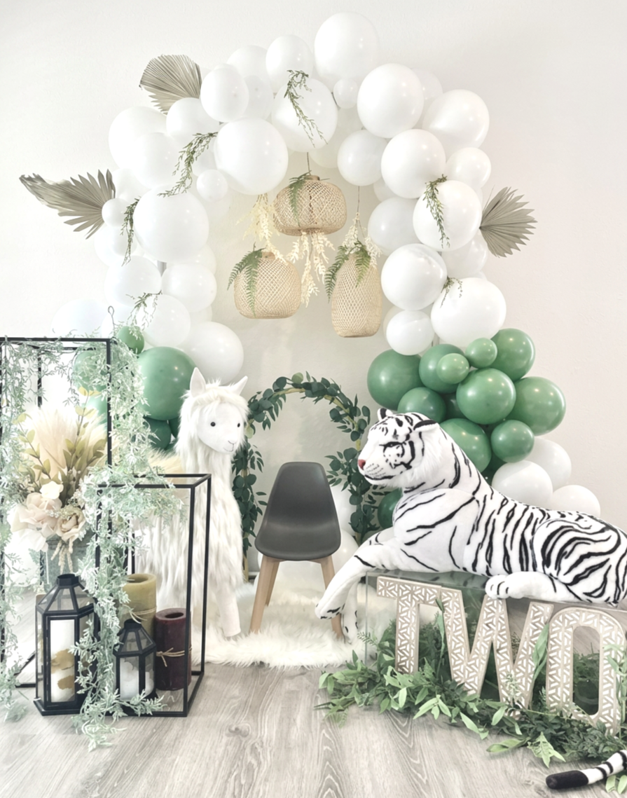 Decorative setup with white and green balloons, hanging woven lanterns, faux plants, a white llama mascot, a stuffed white tiger, an empty gray chair, and a wooden 'TWO' sign, on a light wood floor.