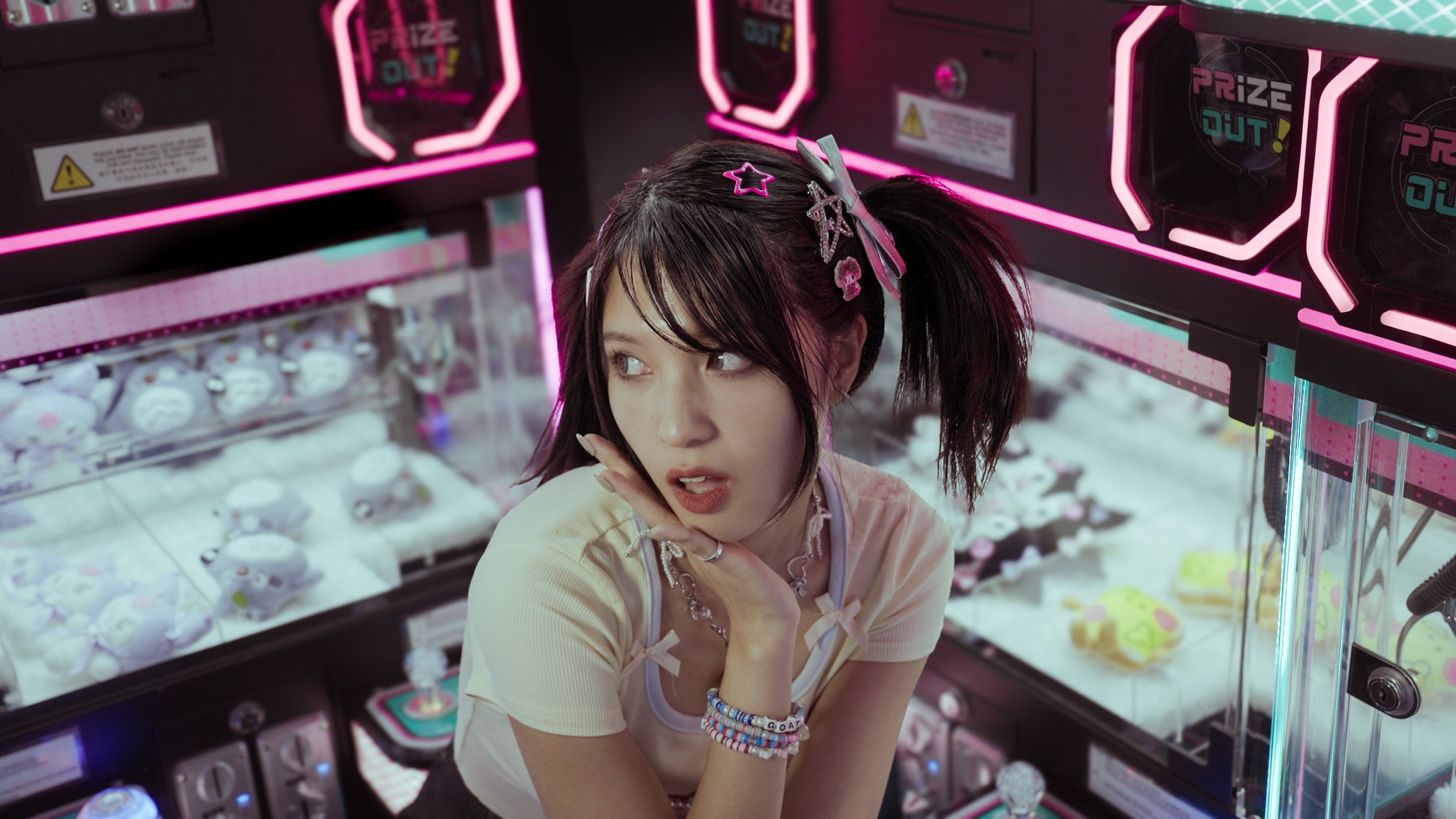 A young woman with brown hair and hair accessories sits in front of a claw machine filled with plush toys, inside an arcade with pink neon lights.