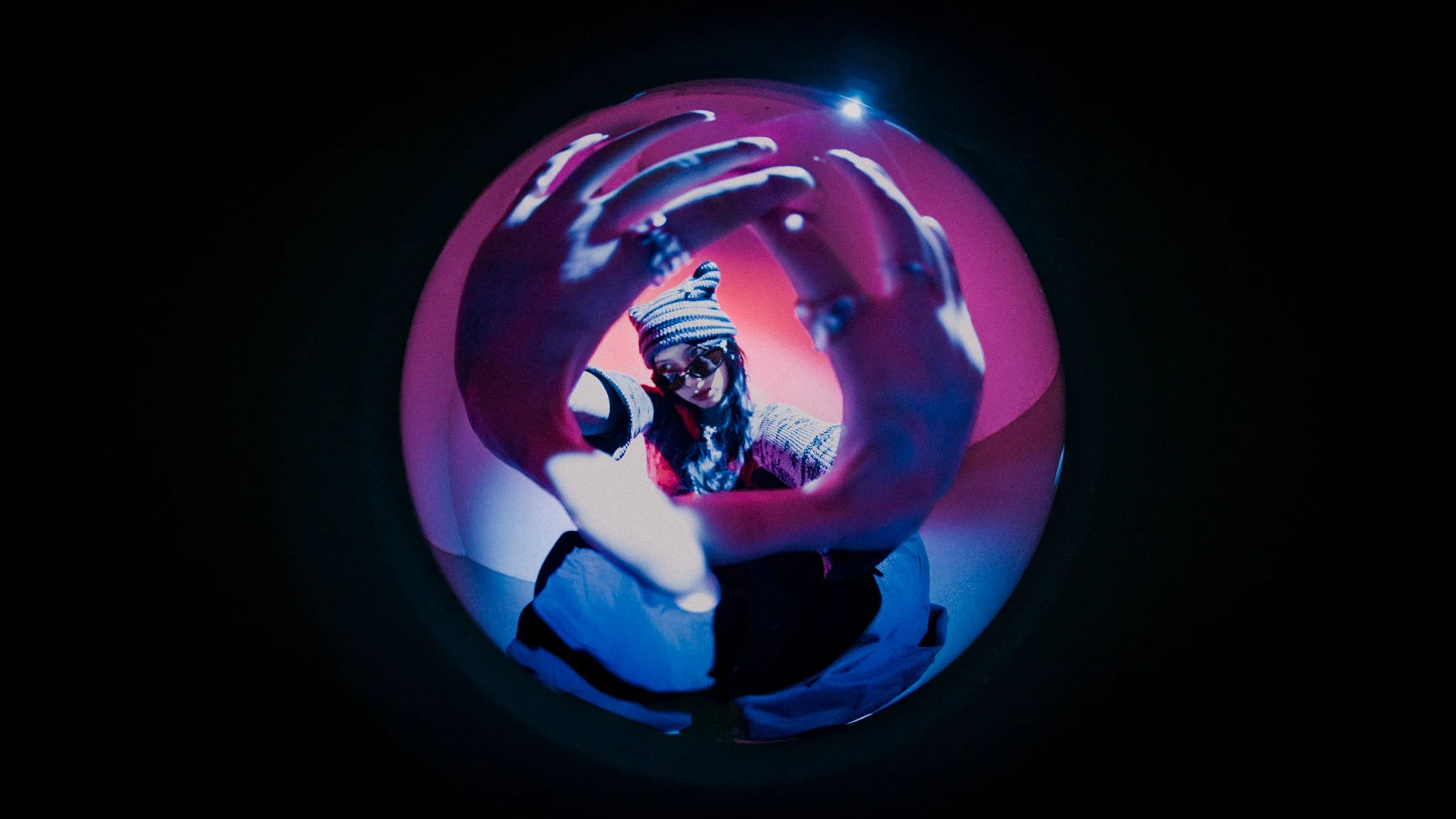 A person with glasses and a striped beanie hat looking into a circular tunnel with pink and purple lighting, creating a fisheye effect.