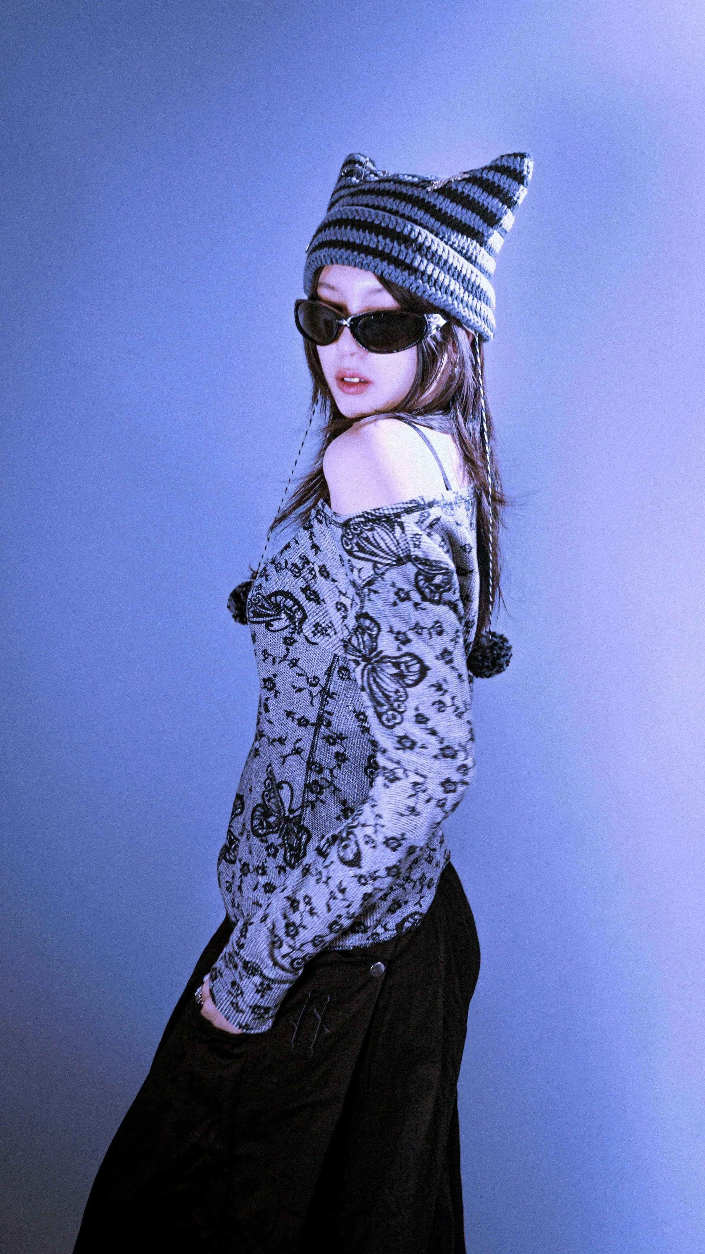 A young woman with wet hair, wearing large black sunglasses, a striped knit hat with pointed ends, a butterfly-patterned sweater, and black pants, standing against a plain light blue background.