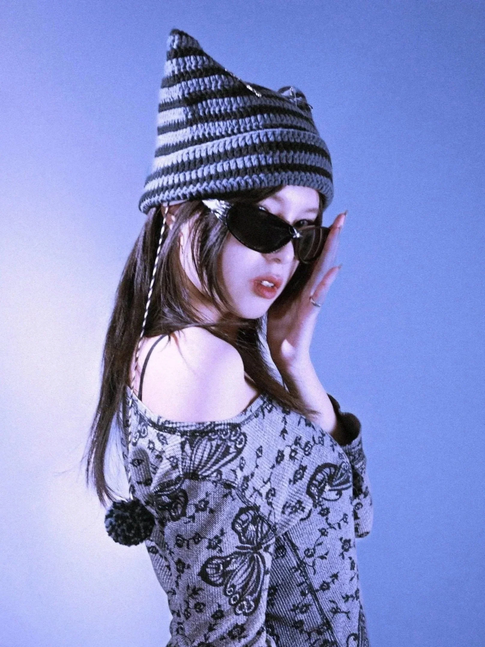Young woman wearing a striped beanie hat, sunglasses, and patterned top, posing and looking over her shoulder against a plain background.