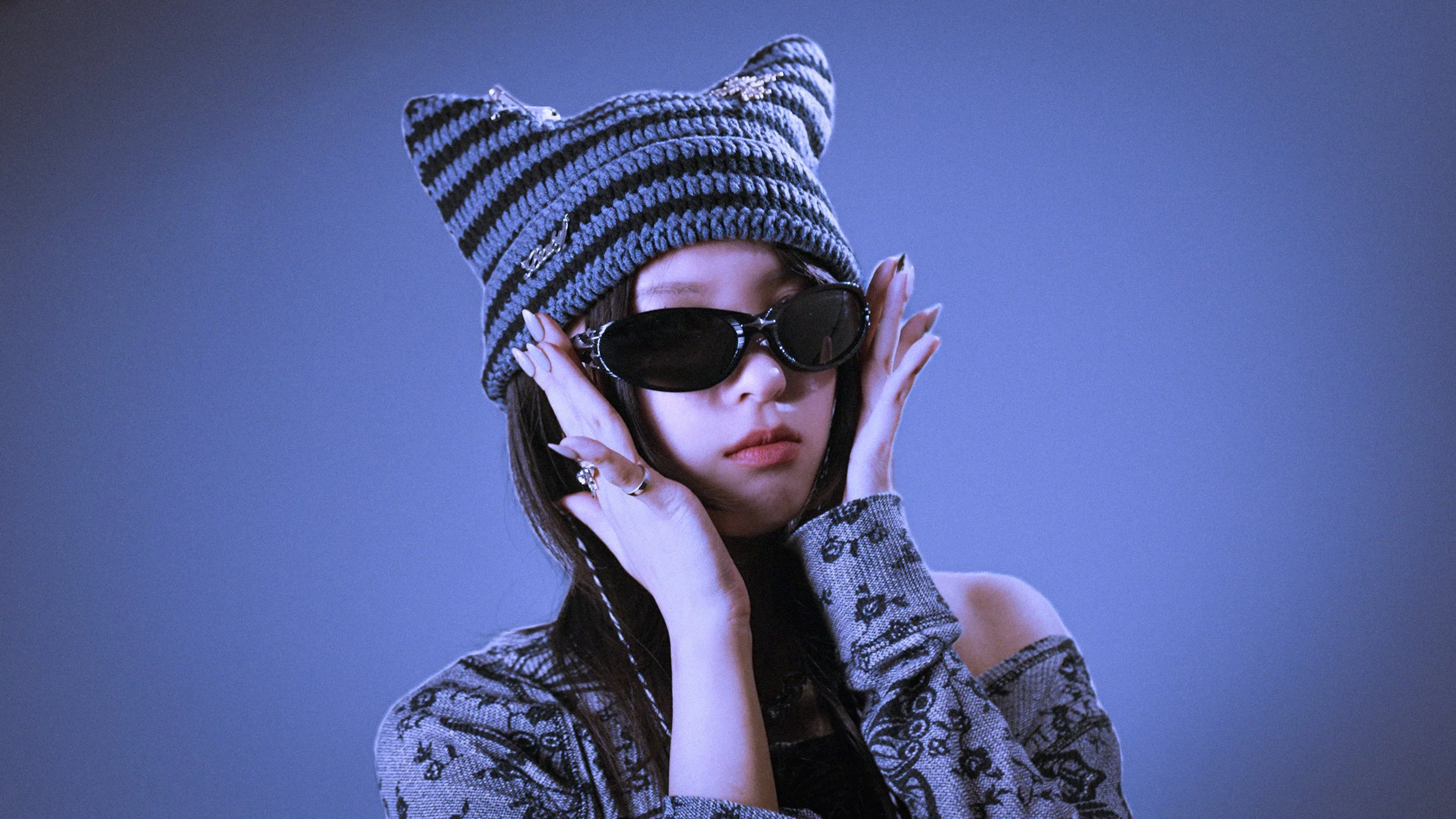 A woman wearing a large, striped knit hat, black sunglasses, and a patterned top, holding her sunglasses with both hands against a plain blue background.