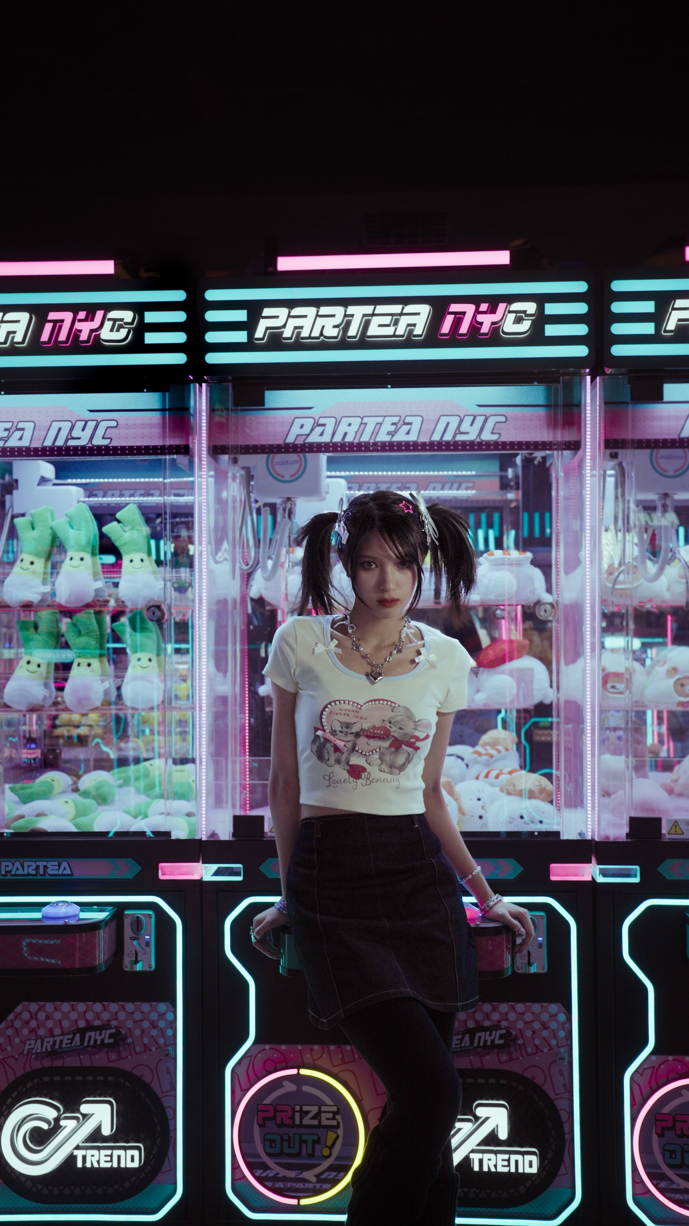 Young woman with pigtails and colorful hair clips posing in front of arcade claw machine filled with plush toys, neon lights, and signs reading 'PARTEA NYC' and 'PRIZE OUT!'