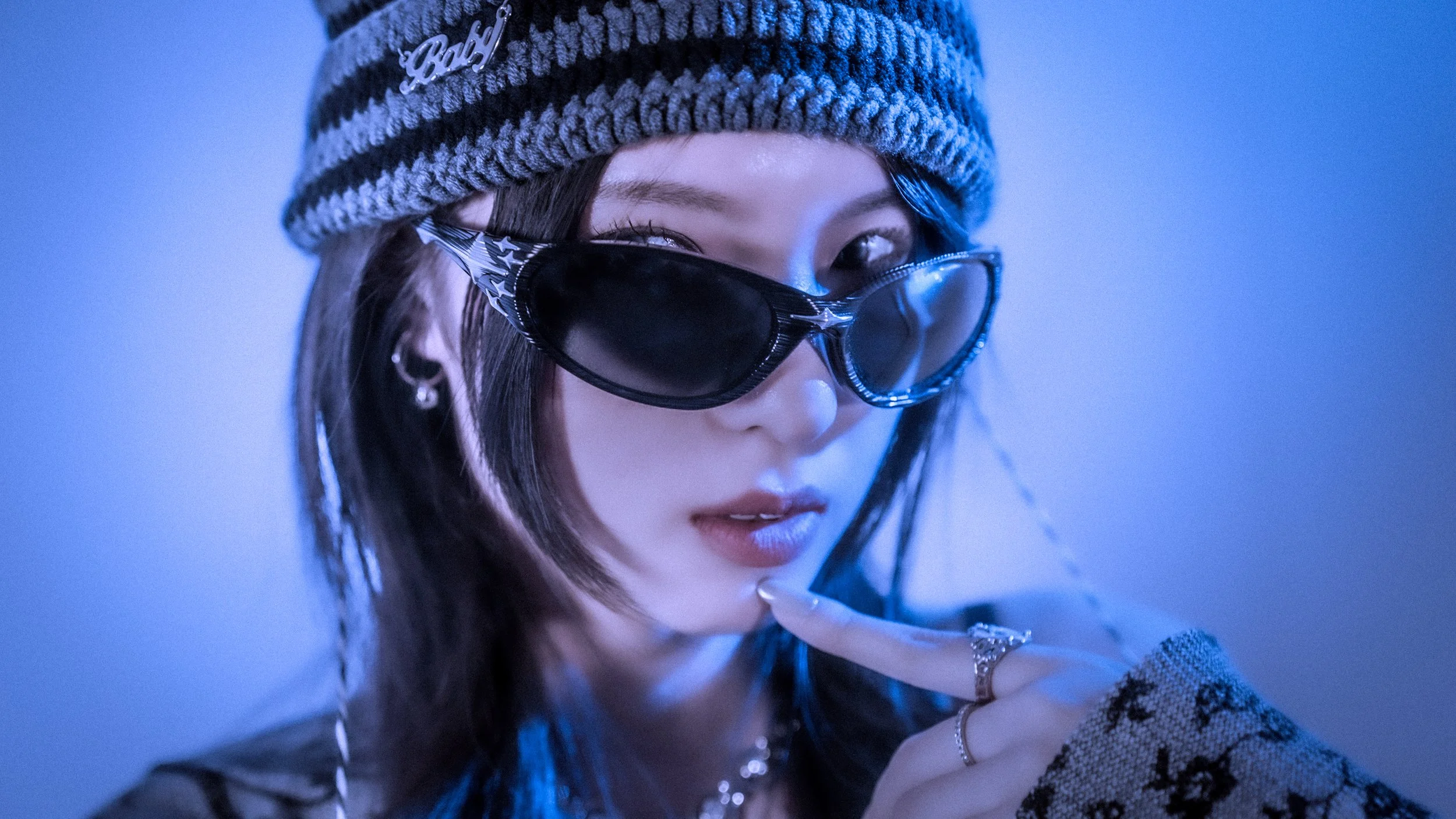 Close-up of a young woman wearing a striped knit beanie hat with a 'Bad' pin and dark sunglasses with star details. She has medium-length dark hair, jewelry including rings, earrings, and a bracelet, and is touching her lips with her finger. The back