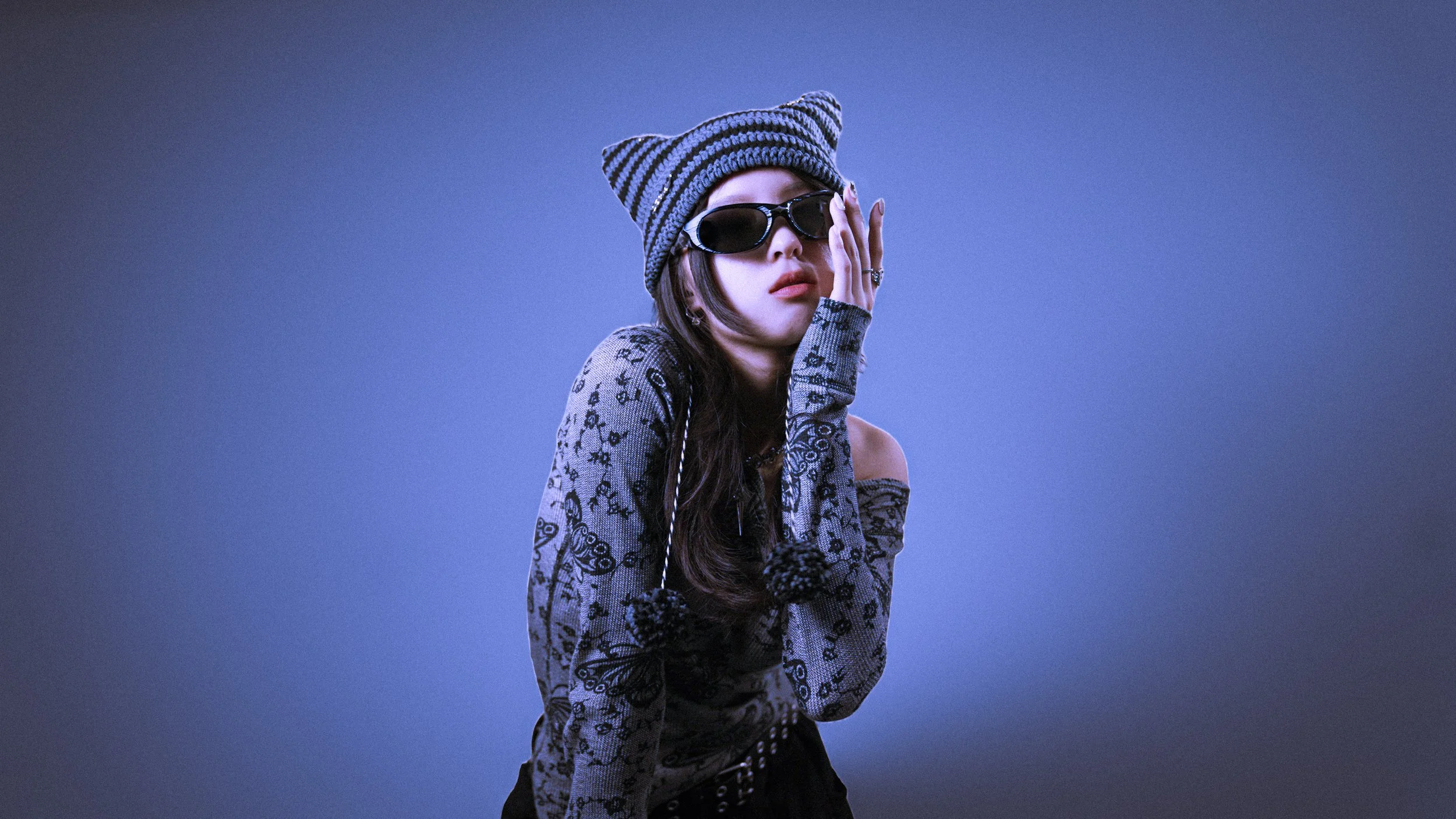 A woman wearing a striped knit beanie, black sunglasses, a long-sleeve top with a floral and butterfly pattern, and black pants poses against a blue background.