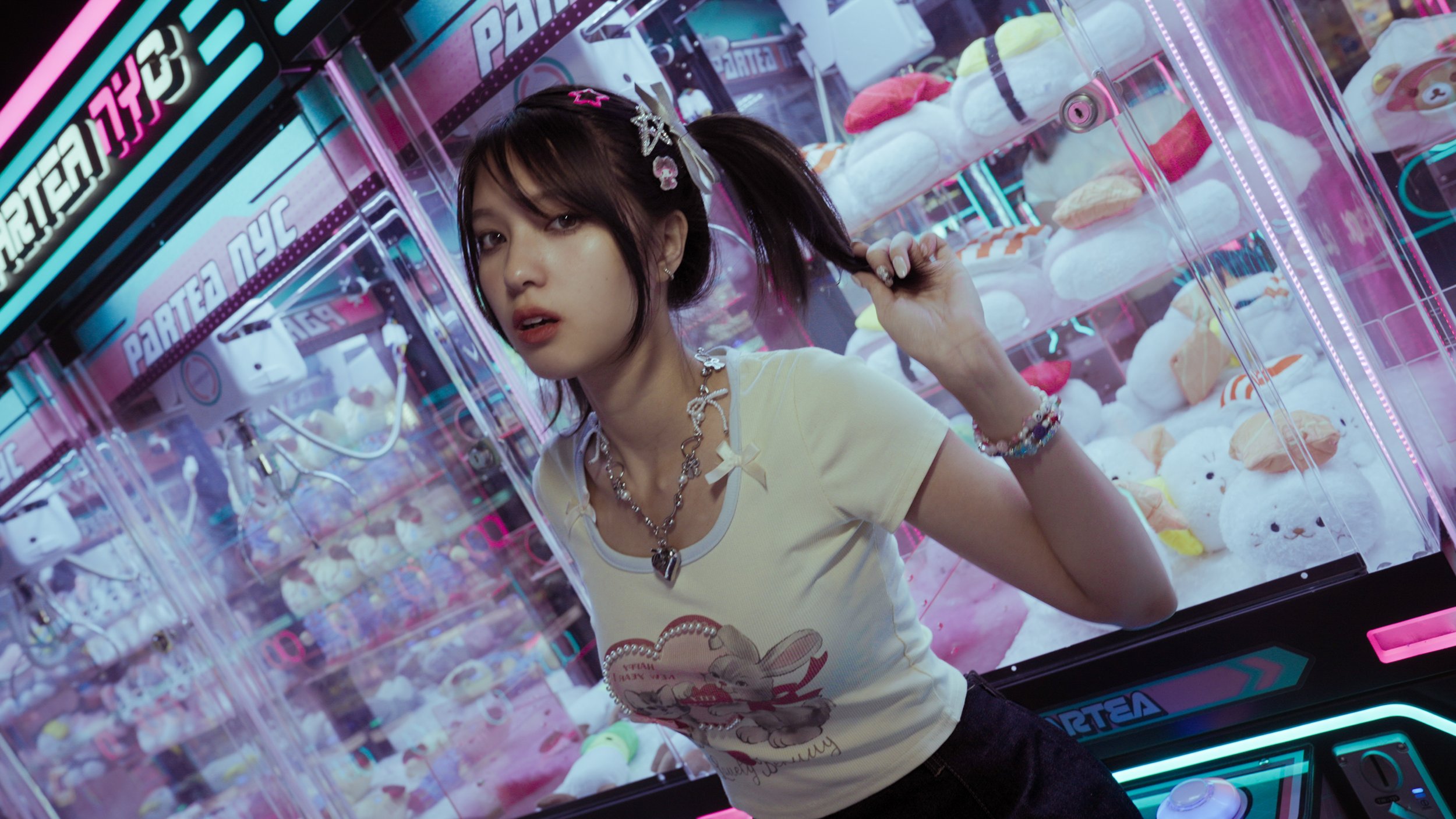 A young woman with dark hair tied in pigtails decorated with colorful hair clips, wearing a yellow T-shirt and layered jewelry, stands in front of a claw machine filled with plush toys at an arcade. Neon lights and colorful signs are visible in the b