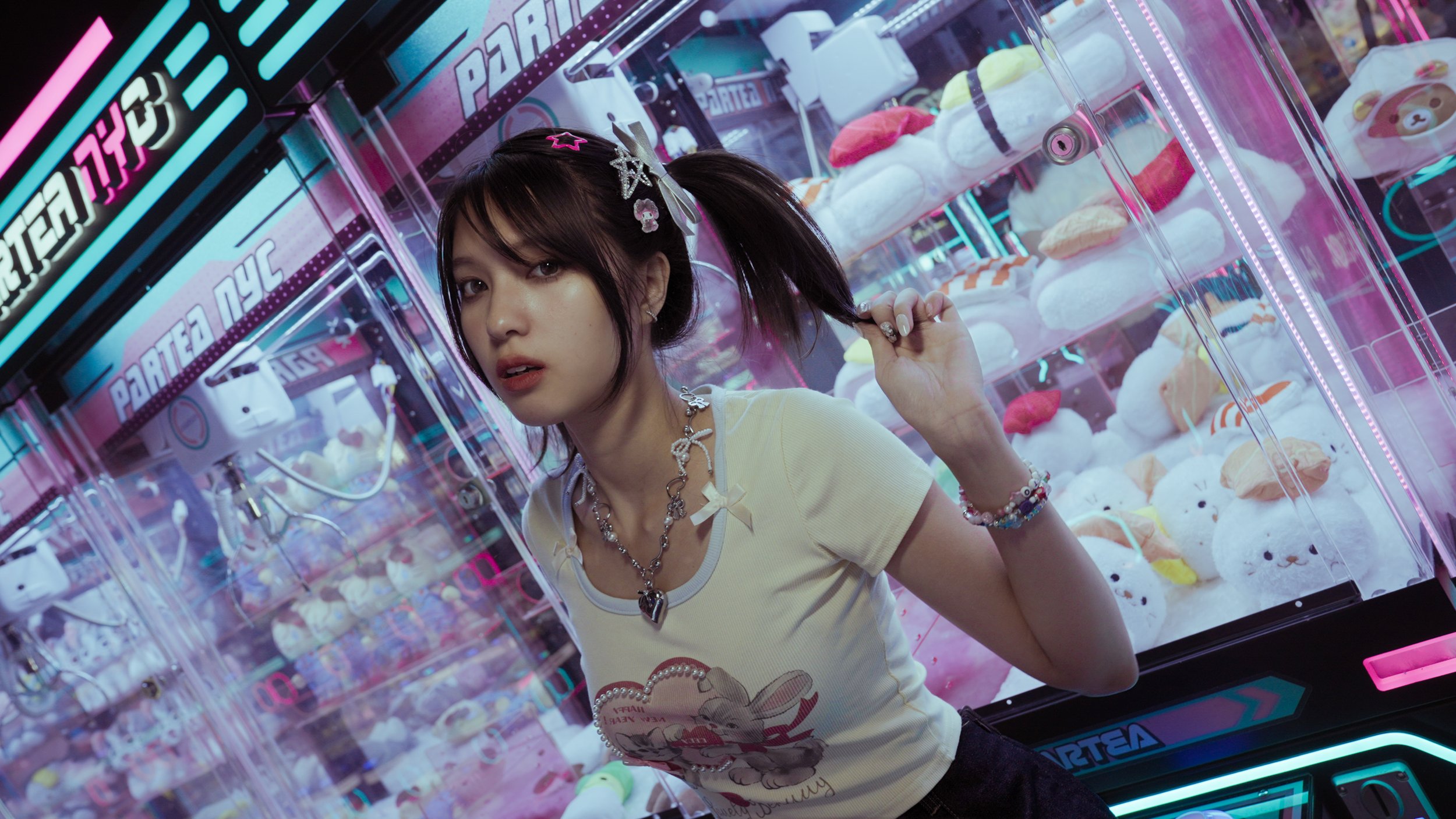 A young woman with dark hair styled in pigtails with colorful hair clips is in front of a claw machine filled with plush toys. She is wearing a light-colored T-shirt with a cartoon graphic, layered jewelry, and colorful bracelets, and is looking at the camera while holding her hair.