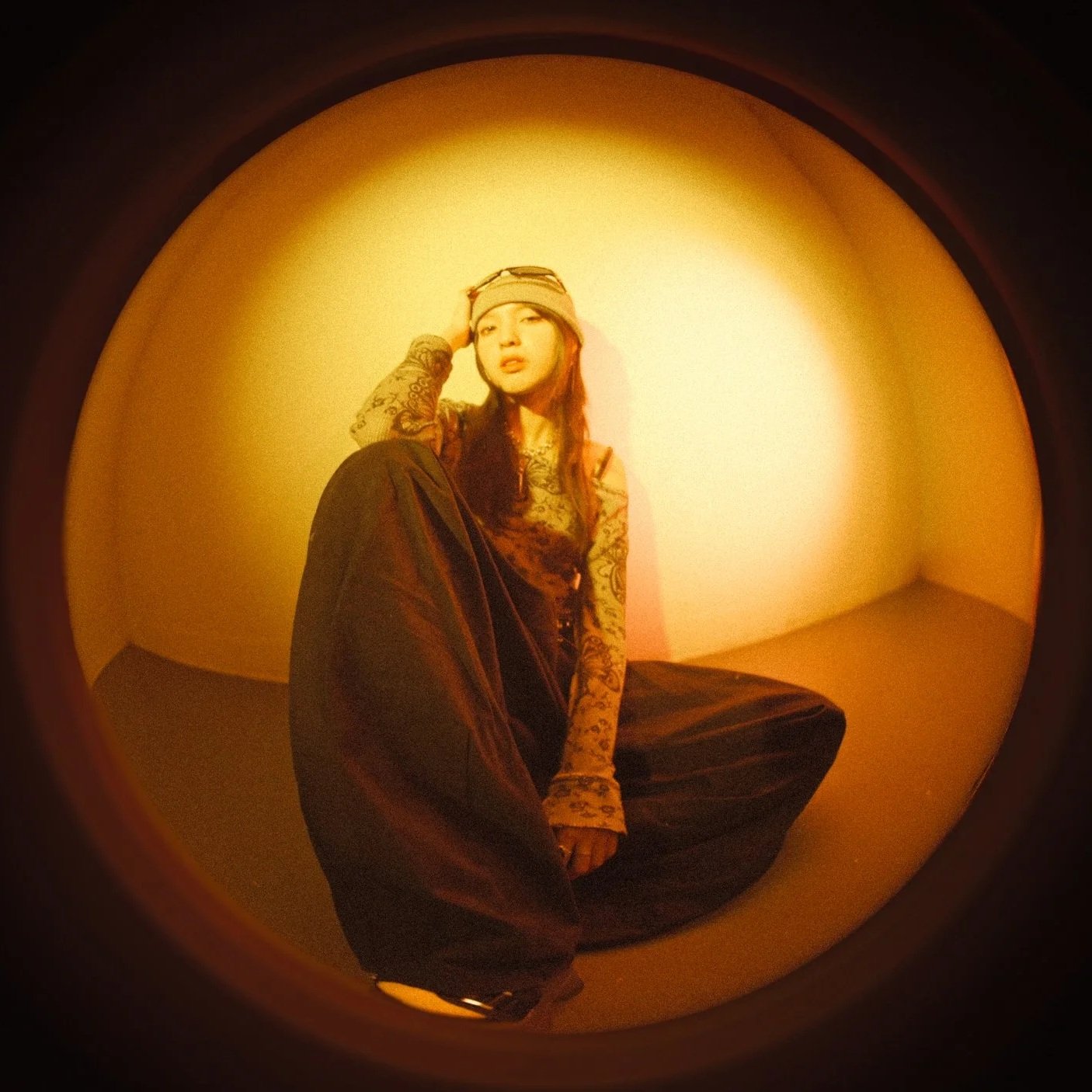 A woman with tattoos, wearing a beanie and long-sleeve shirt, sitting on the floor in a small, warmly lit, empty corner or room, with her arm resting on her knee and looking at the camera.