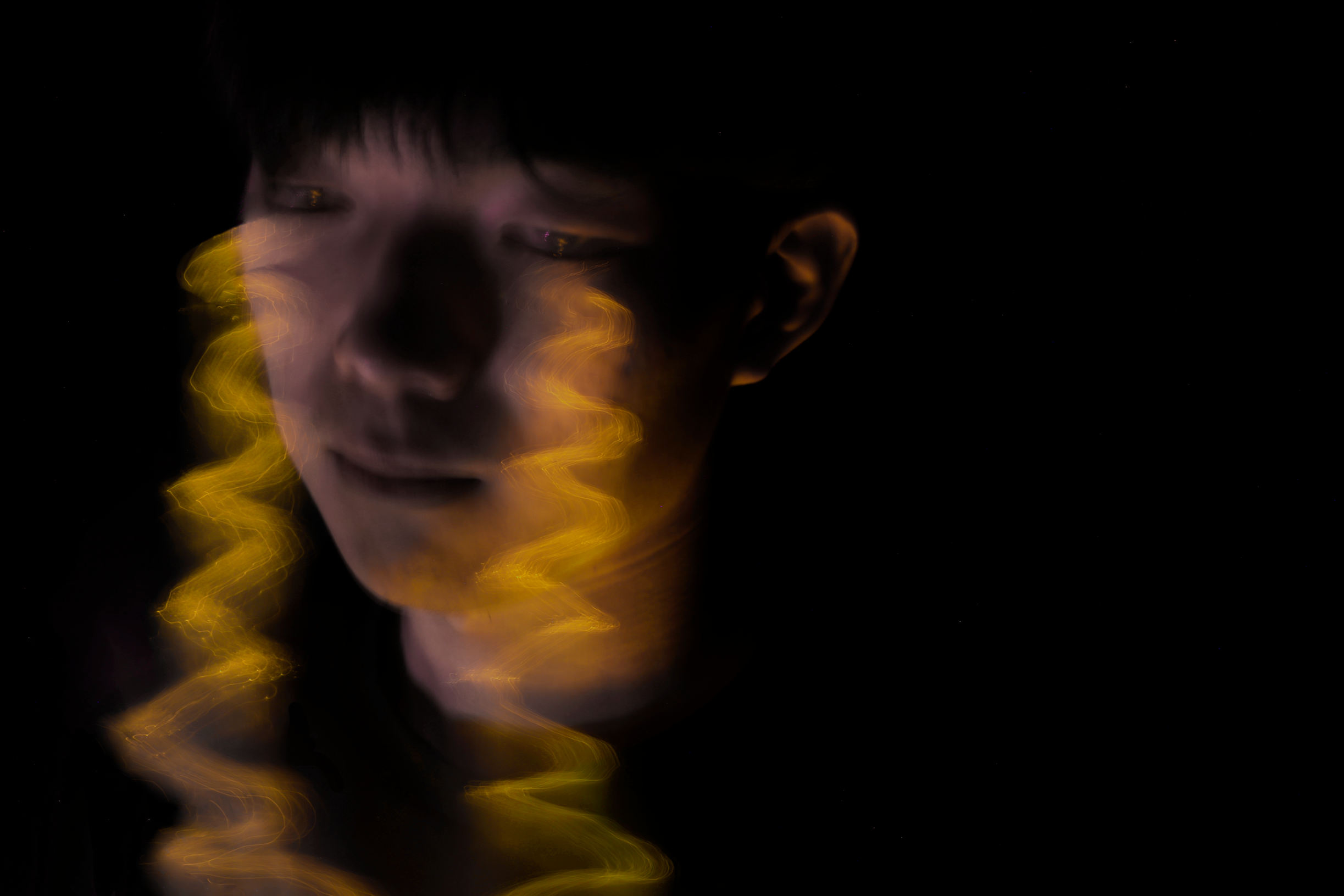 A person with closed eyes and short dark hair, their face partially illuminated by warm, swirling light streaks in a dark background, creating a surreal, abstract portrait.
