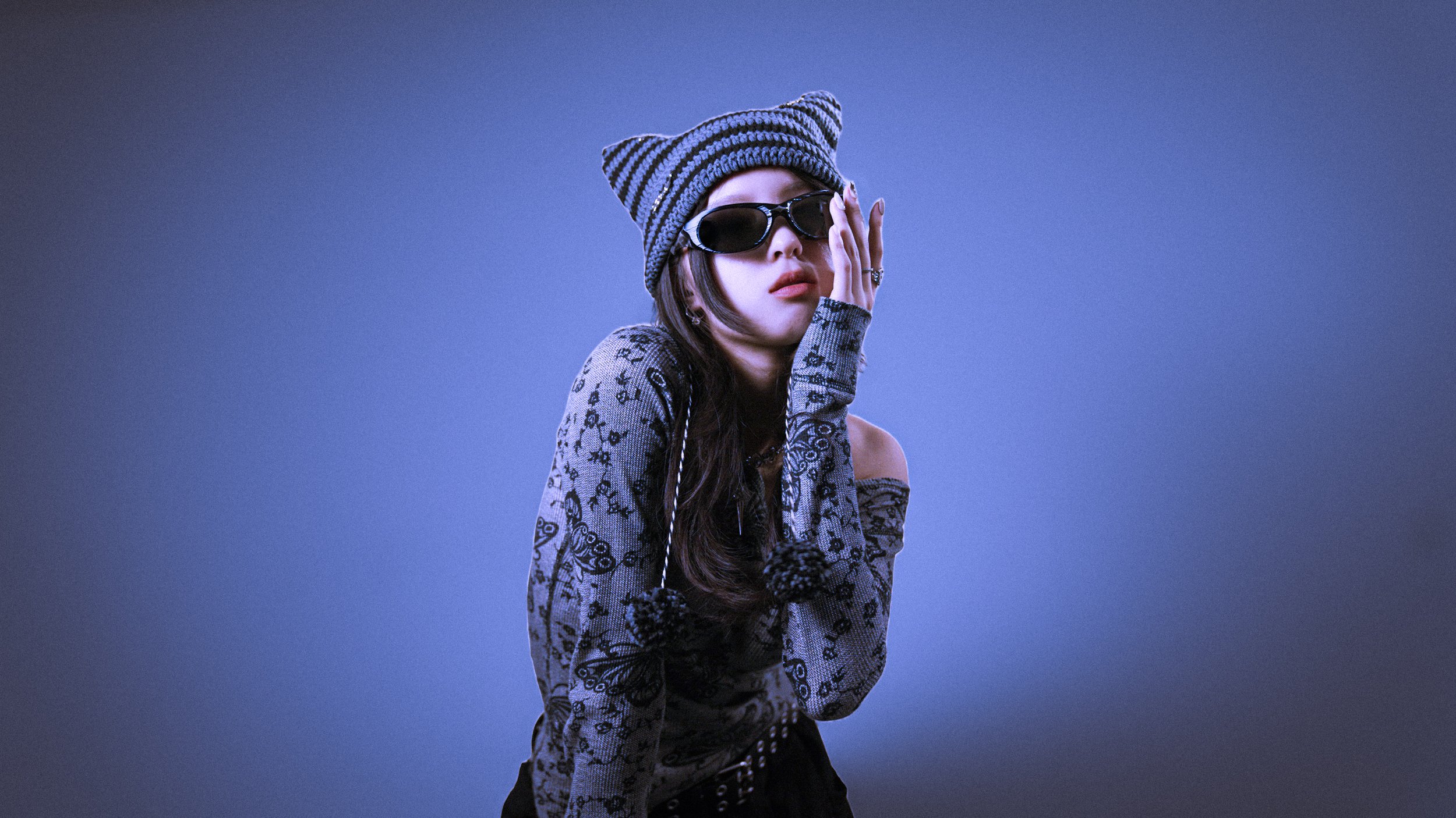 A young woman wearing a black and gray striped beanie, black sunglasses, a long sleeve shirt with black floral and abstract patterns, and black pants, standing against a plain blue background.