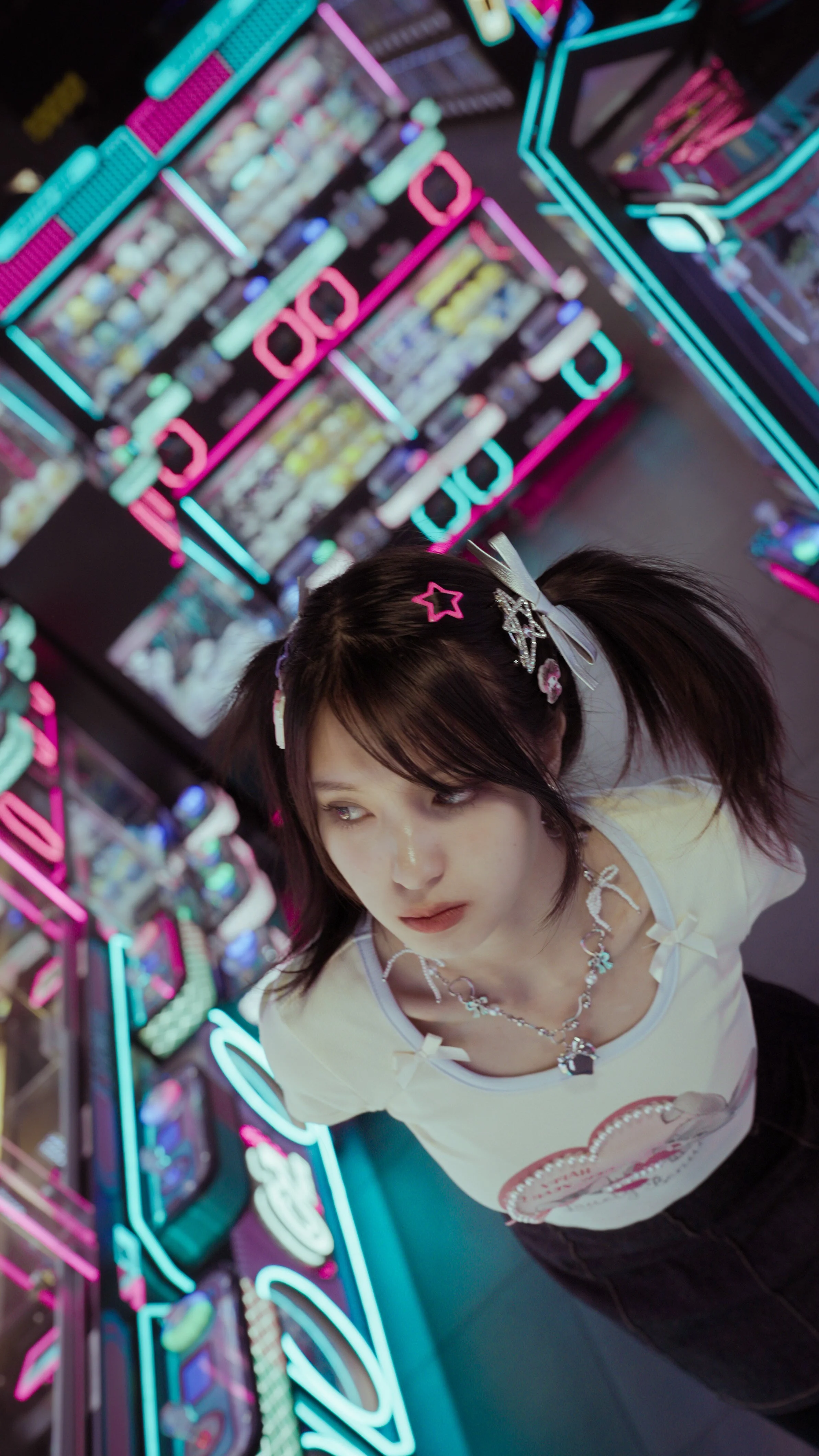 A young woman with dark hair and colorful hair clips standing in front of a bright, neon-lit arcade game machine. She is looking to the side, wearing a light-colored shirt with a graphic and jewelry.