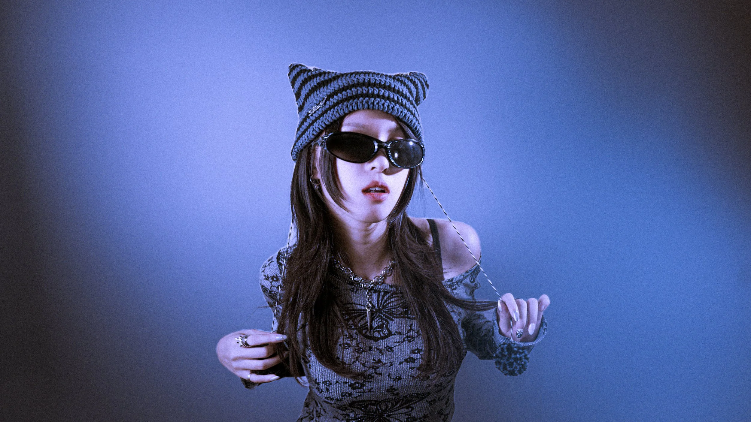 A young woman with long brown hair wearing a black and gray striped knit beanie, black sunglasses, and a patterned top. She has a chain necklace and rings, and is holding a string in her hands against a blue gradient background.