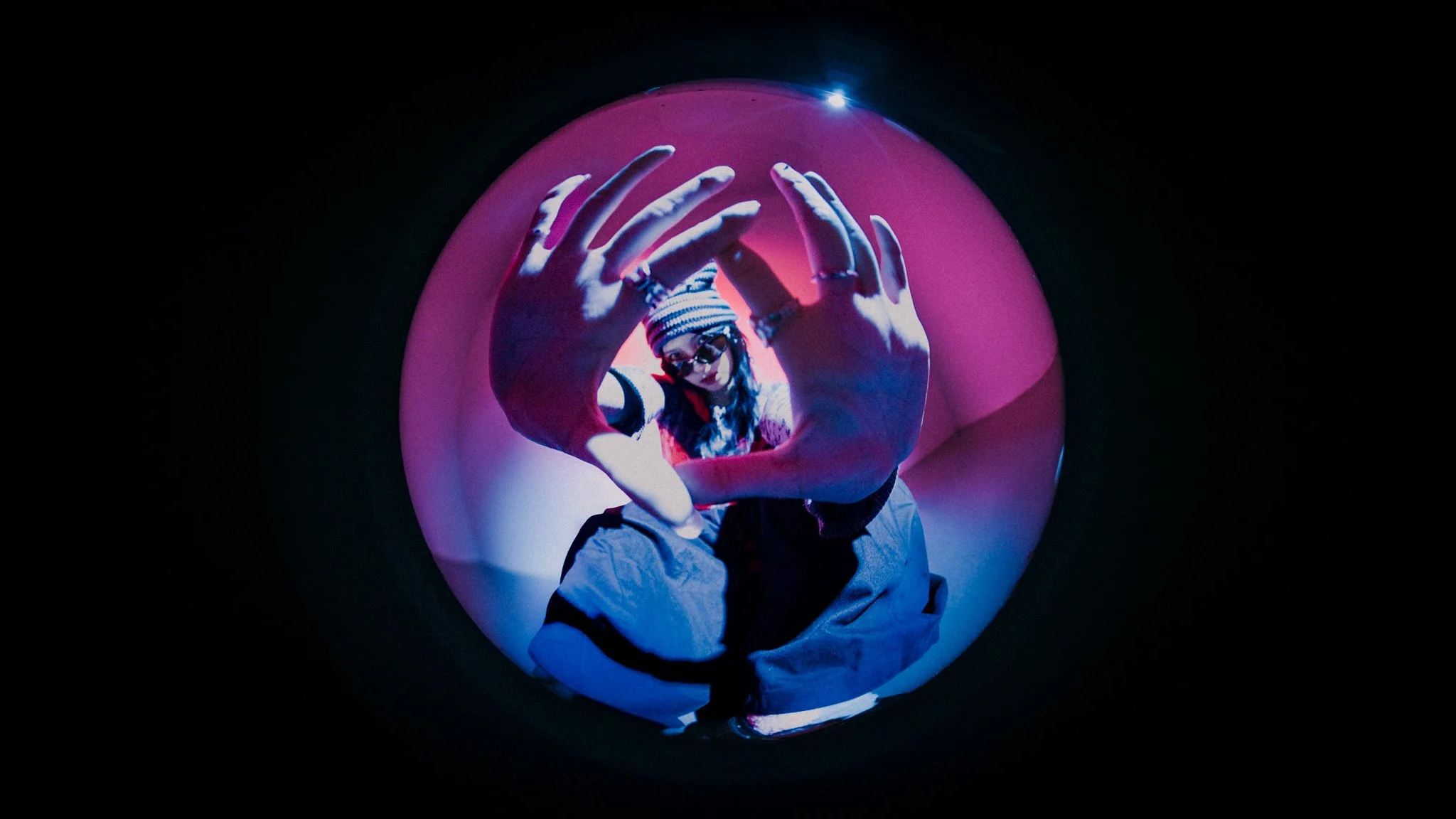 Person wearing striped shirt, glasses, and beanie hat, reaching out with hands in front of them inside a pink, spherical tunnel with dark surroundings.
