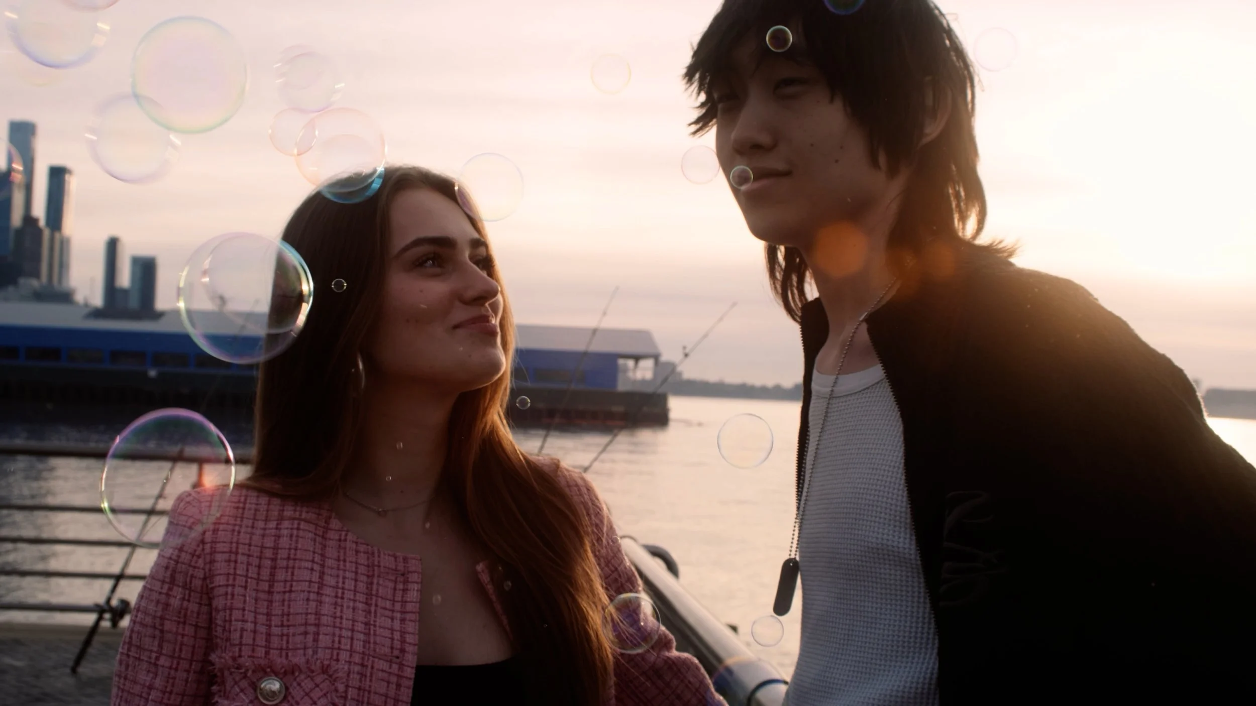 A young woman and a young man stand close to each other by the water during sunset, with bubbles floating around them.