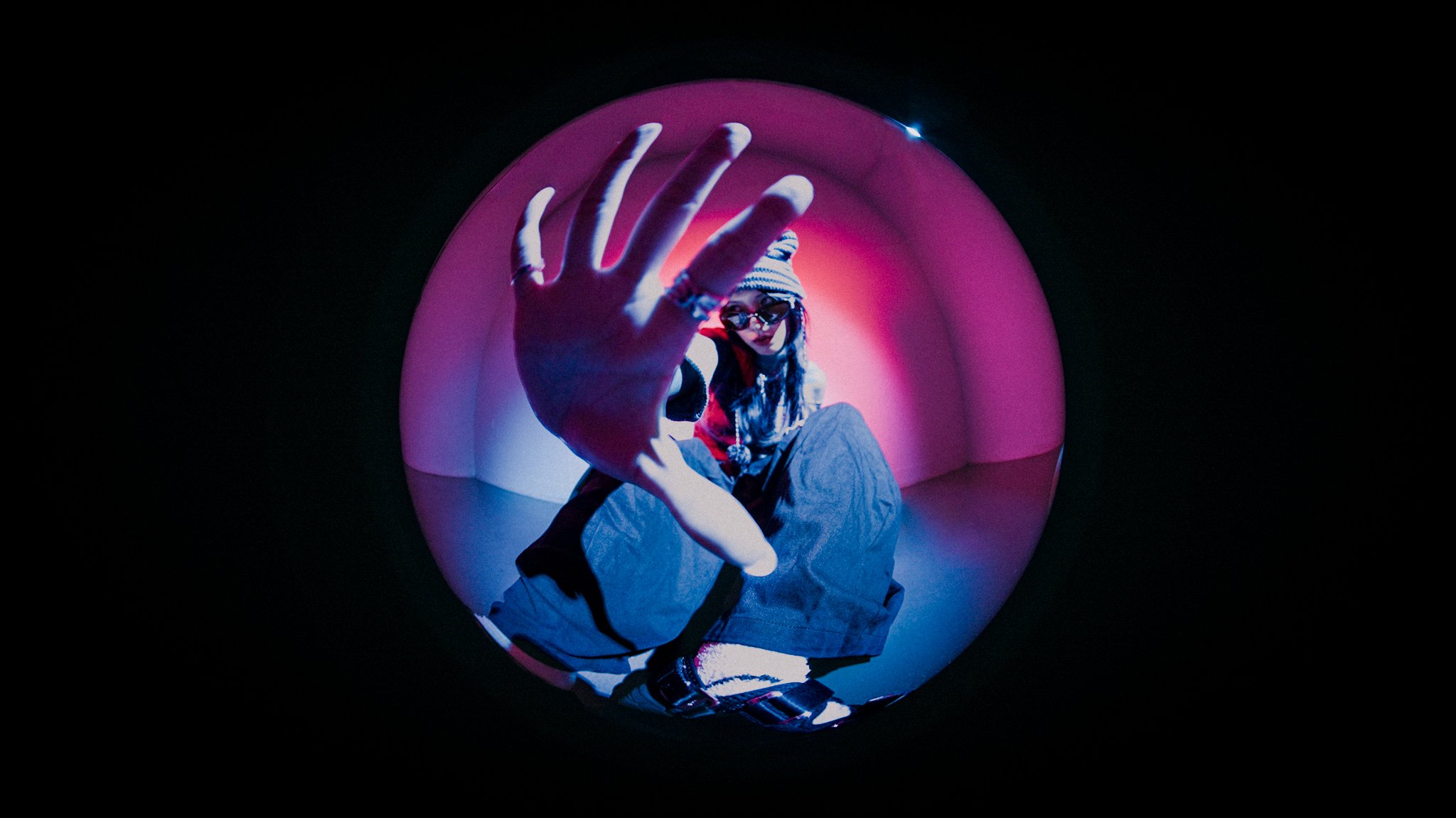 Person wearing sunglasses and a beanie, crouching inside a reflective sphere with pink and purple lighting, reaching out with an open hand.