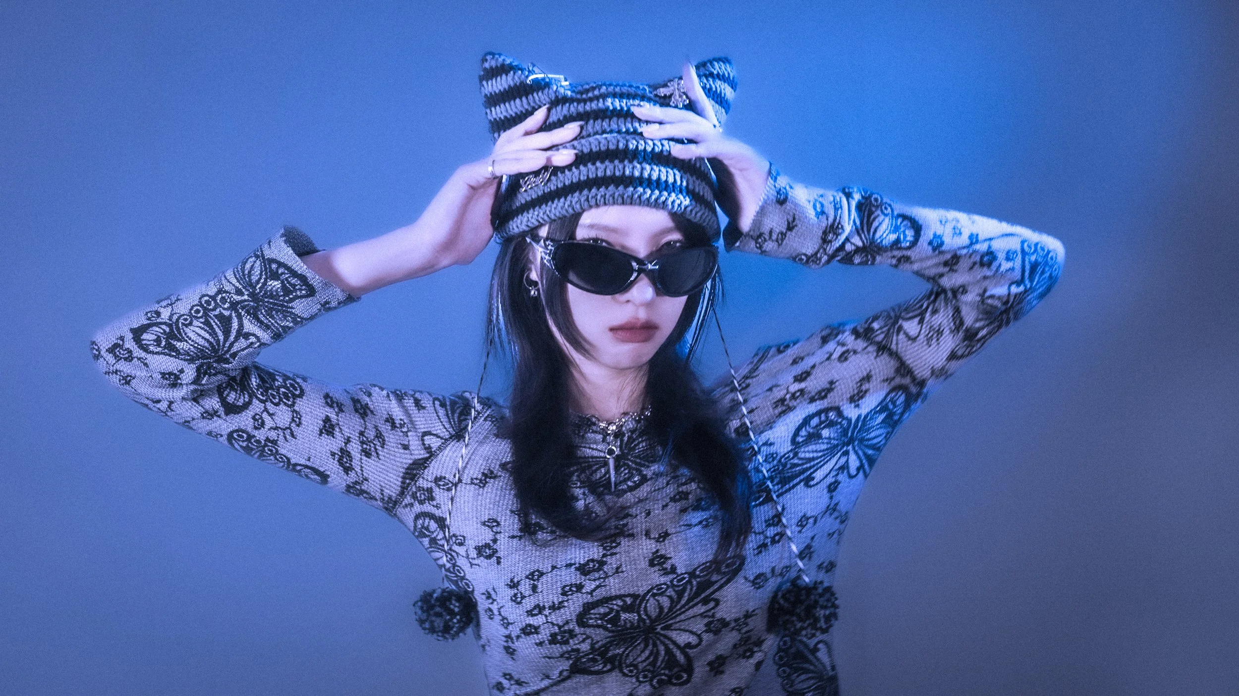 A woman wearing large black sunglasses and a patterned long-sleeve top with butterflies and flowers, holding a striped knit beanie on her head against a blue background.