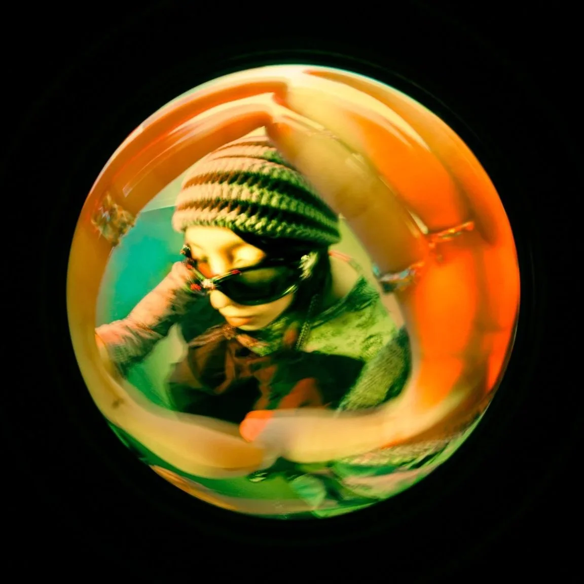 A person wearing a striped beanie, sunglasses, and a green jacket taking a selfie in a reflective, multicolored spherical surface.