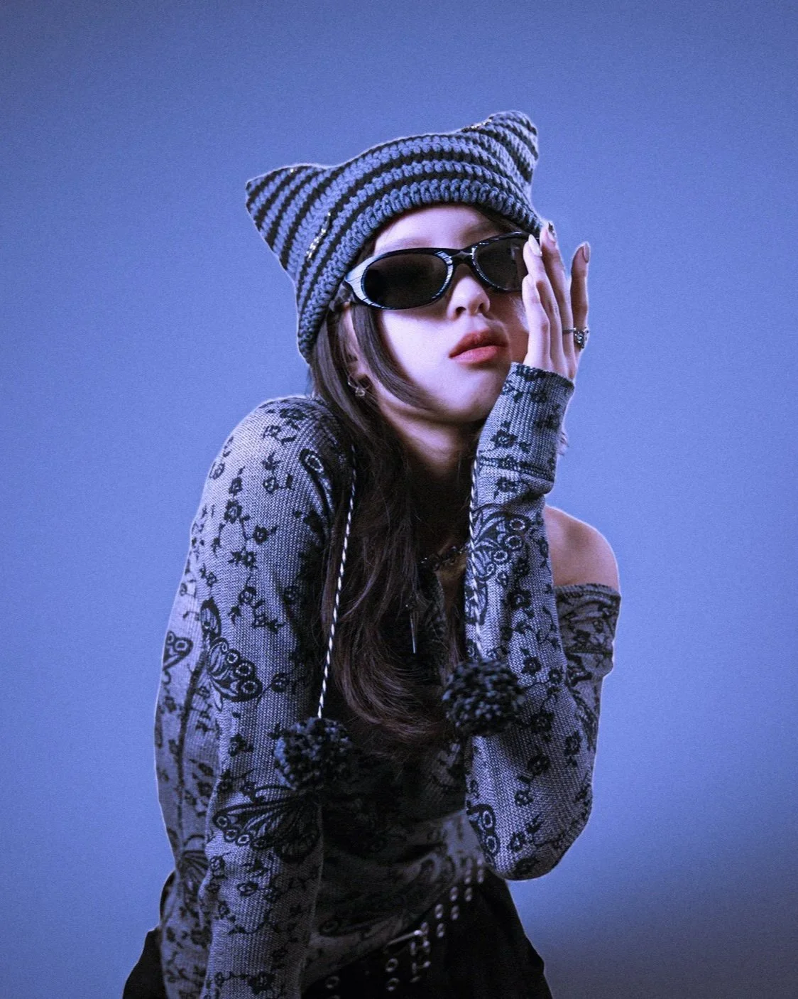 A young woman with long dark hair wearing a striped knit beanie, black sunglasses, and a gray floral patterned top, stylishly posing with her hand on her face against a plain blue background.