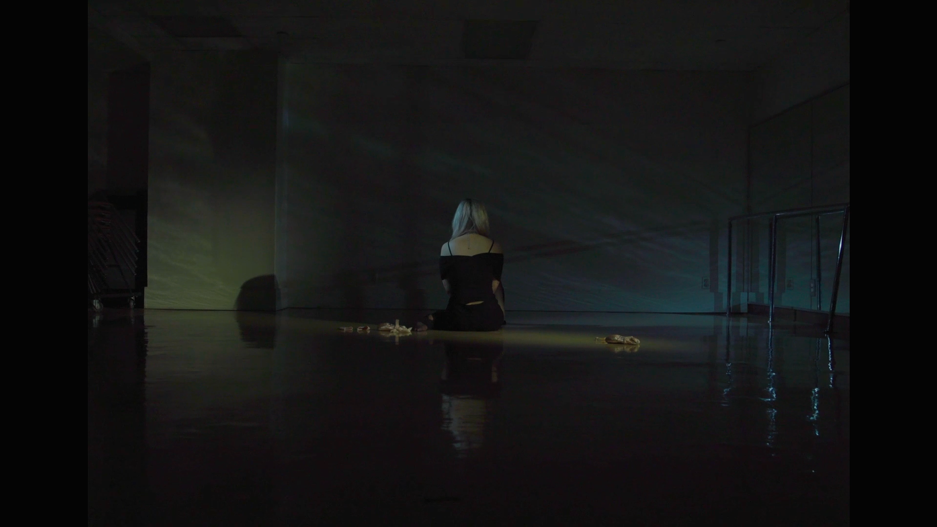A woman with blonde hair sitting cross-legged on the floor in a dimly lit room, facing away from the camera. She is wearing a black off-the-shoulder top with cut-outs. The room has dark walls, and there are a few objects around her, including some shoes. The lighting creates a soft, moody atmosphere with light reflections on the floor.