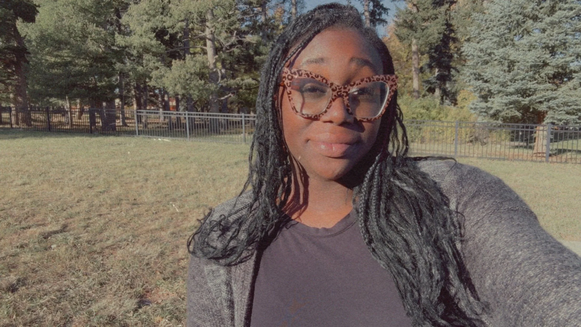 A woman outdoors with black and gray braided hair, wearing large, leopard print glasses, a dark gray shirt, and a gray jacket. She is smiling and standing on grass with trees and a metal fence in the background.