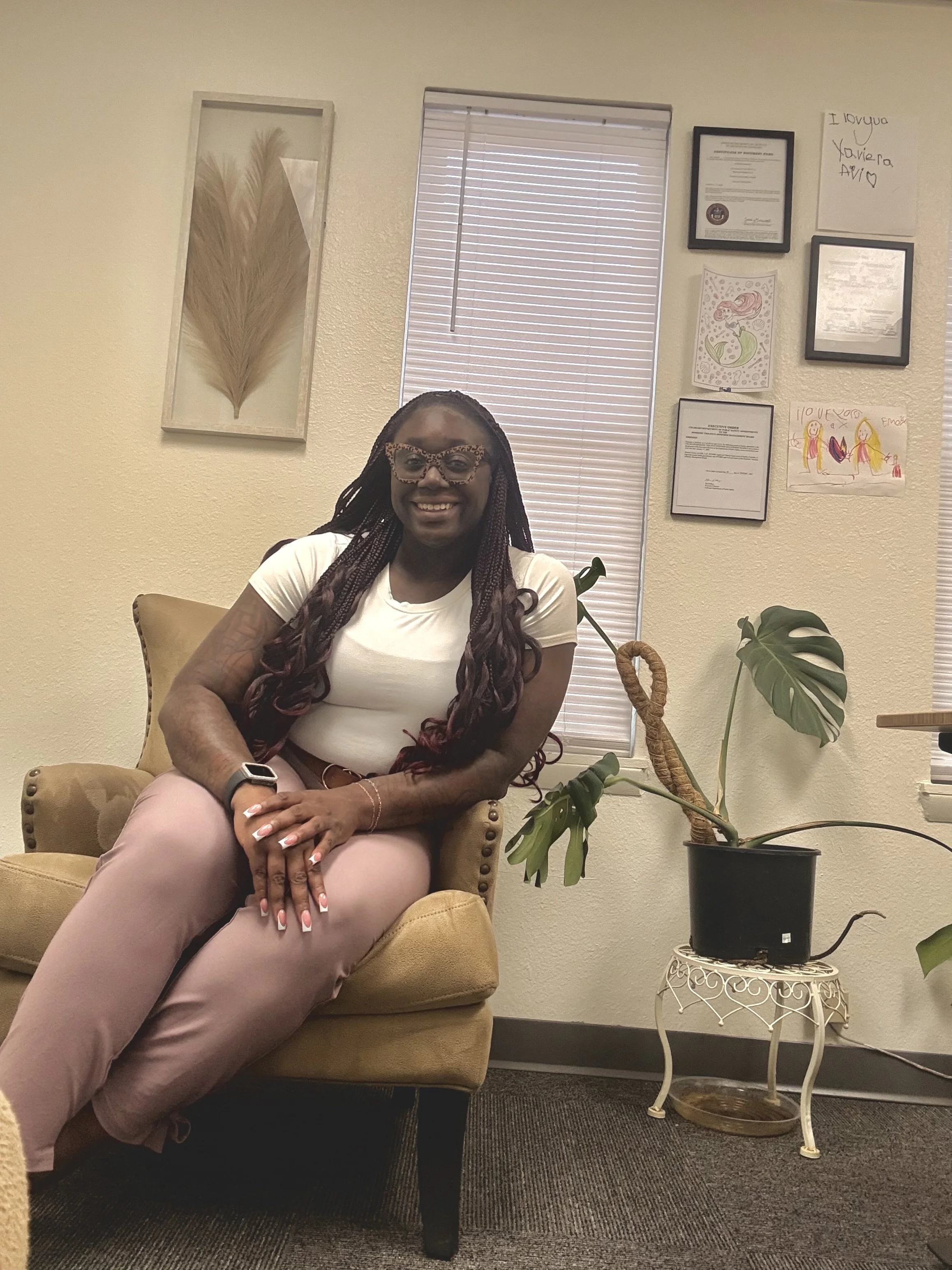 A woman with dark braided hair and glasses sitting on a beige armchair in a room with framed certificates, drawings, and a leaf artwork on the wall, a window with blinds behind her, and a large potted plant beside her.