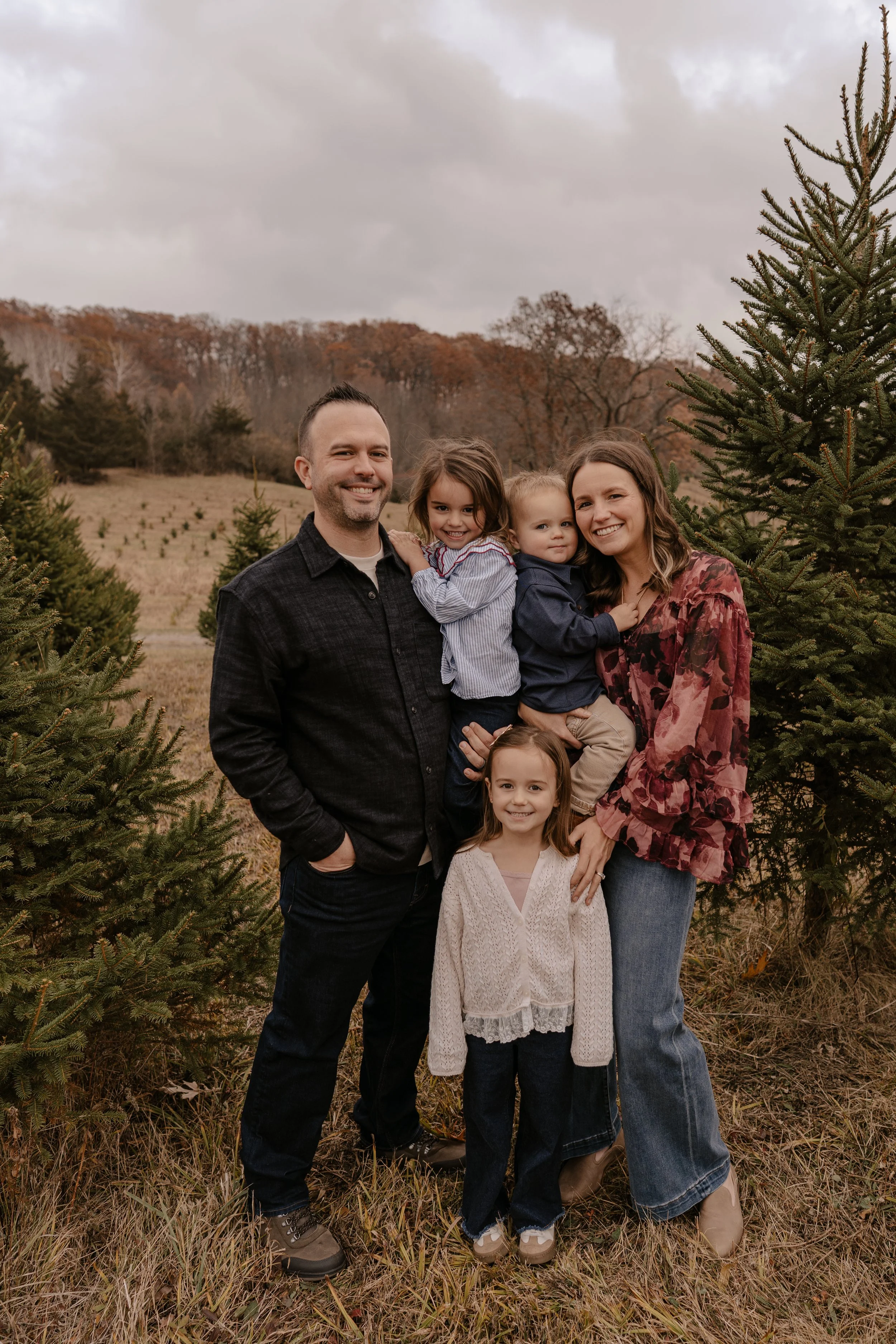 Family of six standing outdoors among Christmas trees, smiling, with a cloudy sky and autumn trees in the background.