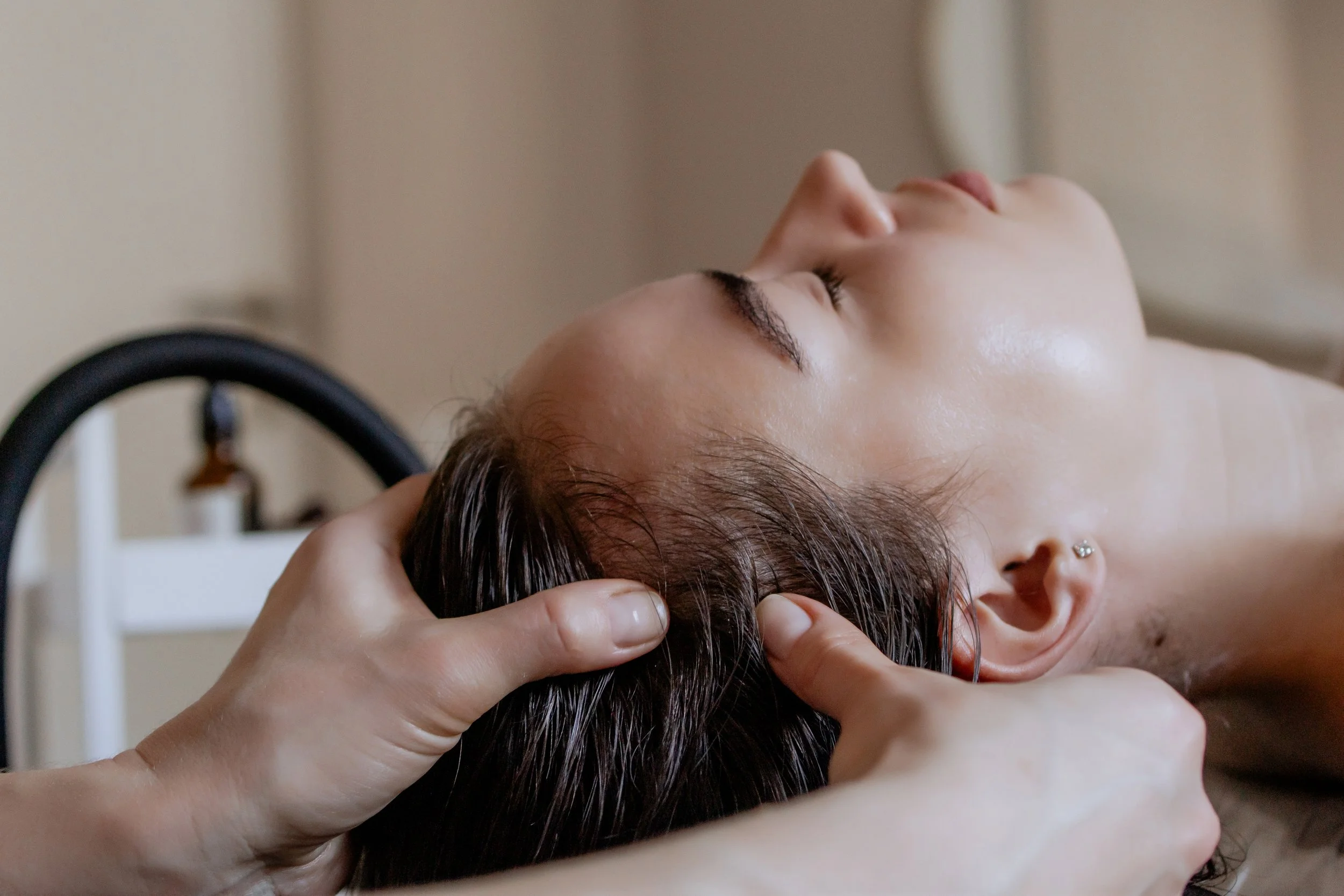A woman receiving a massage with her eyes closed, lying on her back, with one hand supporting her head and the other on her forehead, in a serene indoor setting.