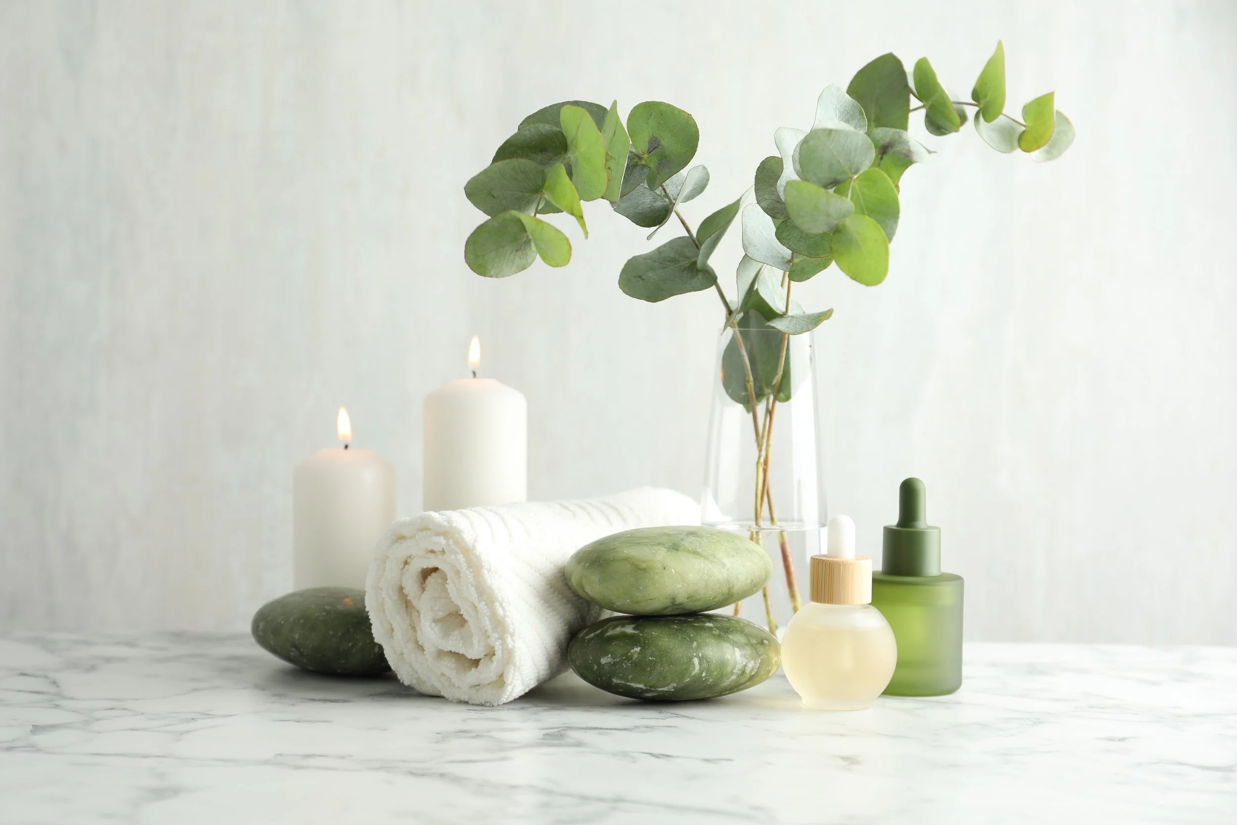 Spa setting with rolled white towels, green stones, candles, and green glass bottles arranged on a marble surface with a tall glass vase containing eucalyptus leaves in the background.