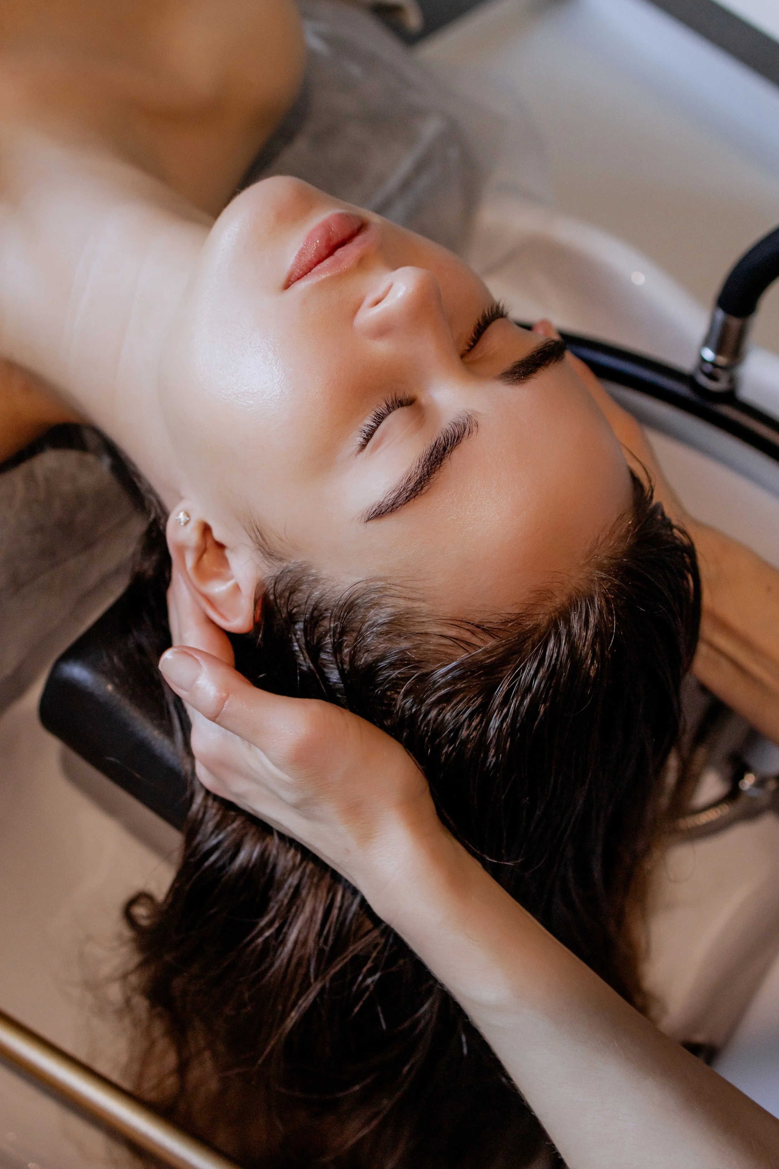A woman with dark brown hair lying on a massage or spa bed with her eyes closed, receiving a head or scalp massage from a person with light skin, in a spa or wellness setting.