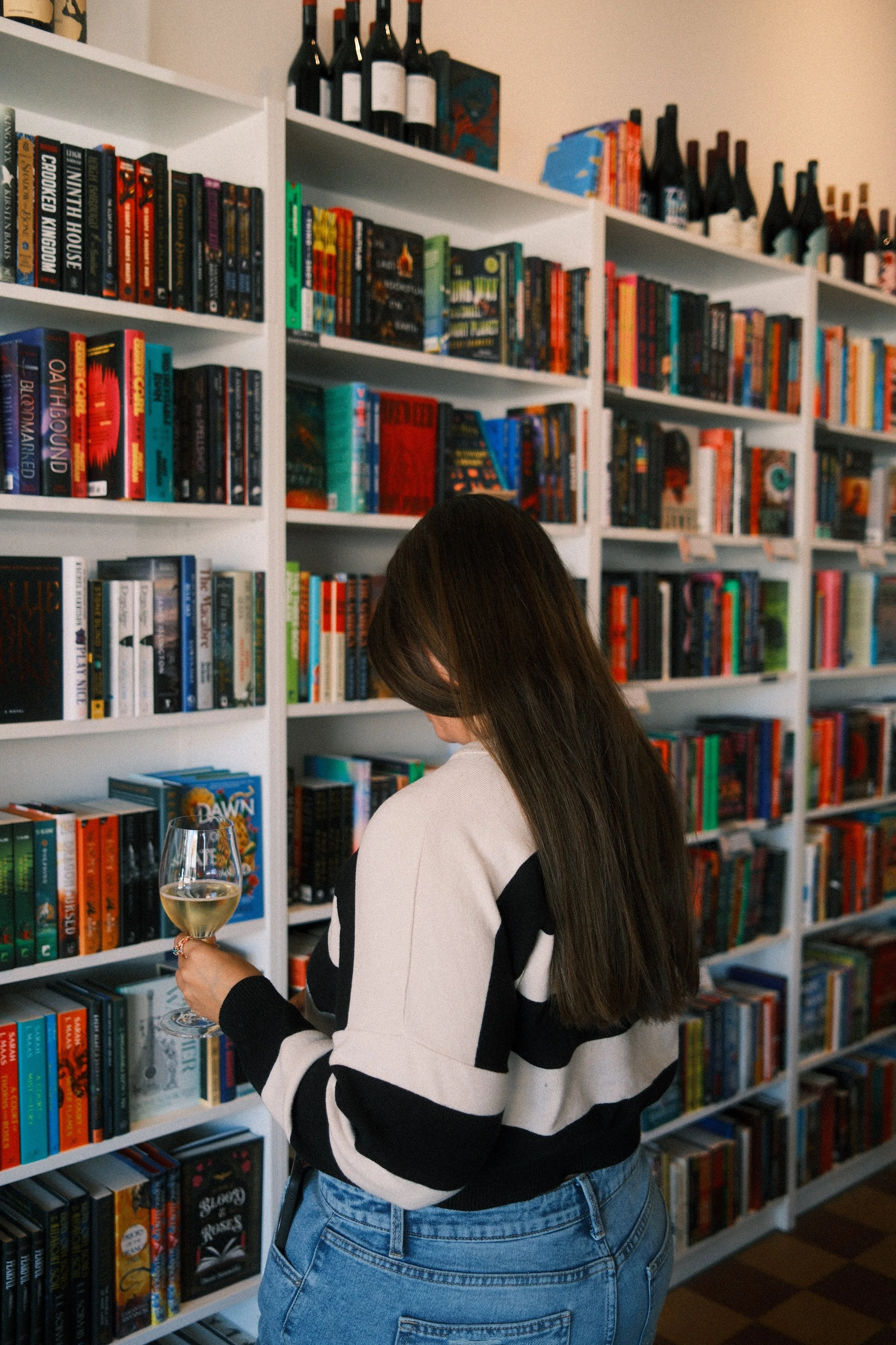 A woman with long brown hair wearing a black and white striped sweater and blue jeans, holding a glass of white wine, standing in front of a bookshelf filled with colorful books.