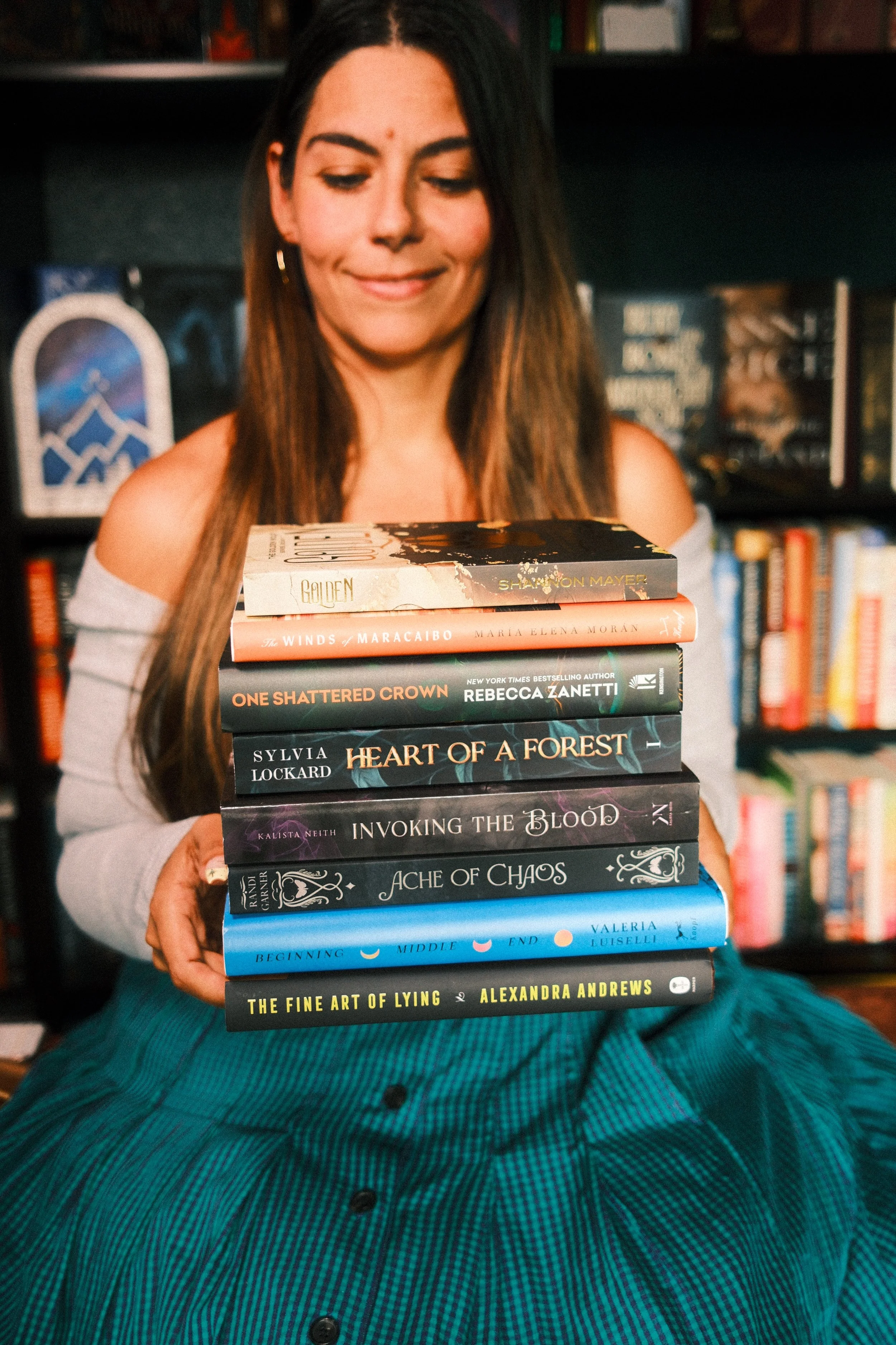 A woman sitting on a teal plaid blanket holding a stack of ten books in a bookstore.