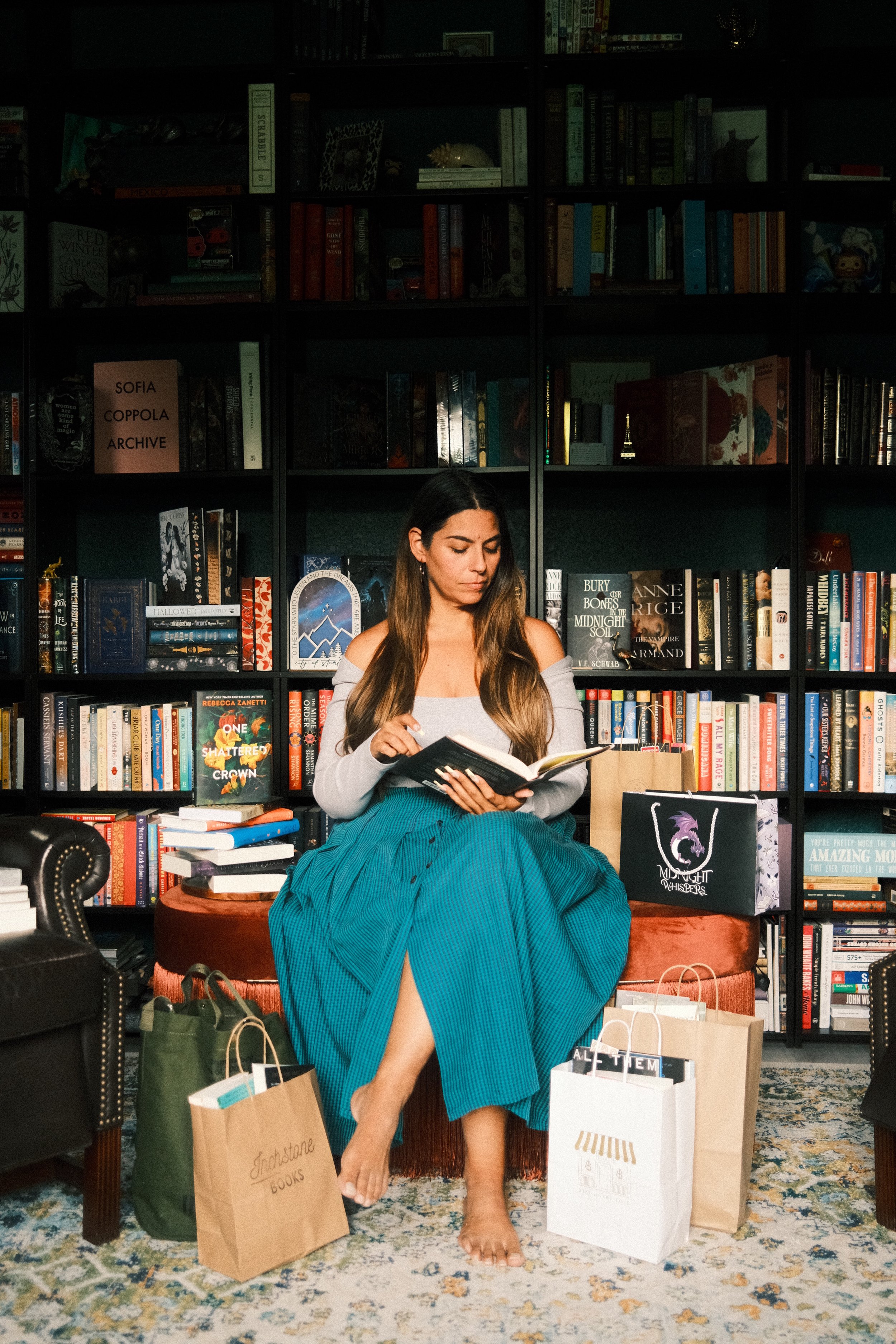 A woman with long brown hair sits on a red ottoman, reading a book. She is barefoot and wearing an off-the-shoulder gray top and a long blue skirt. She is surrounded by shopping bags on the floor, in front of a large black bookshelf filled with books and decorative items. The room has a patterned rug, and there is a black leather chair partially visible on the left.
