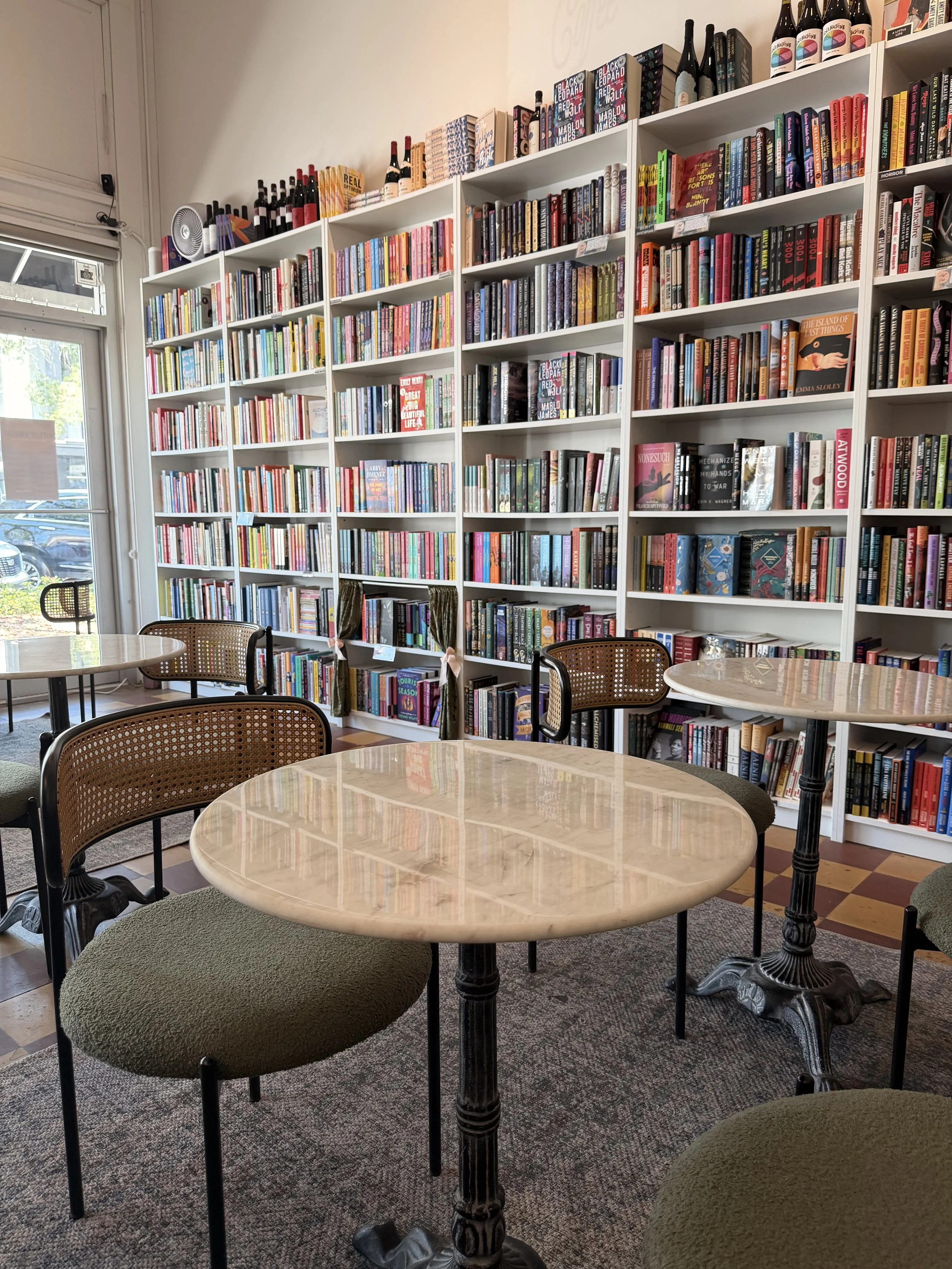 Interior view of a cozy coffee shop or bookstore with several round tables and chairs. Large white bookshelves filled with colorful books and decorative bottles line the wall. Large windows let in natural light, illuminating the space.