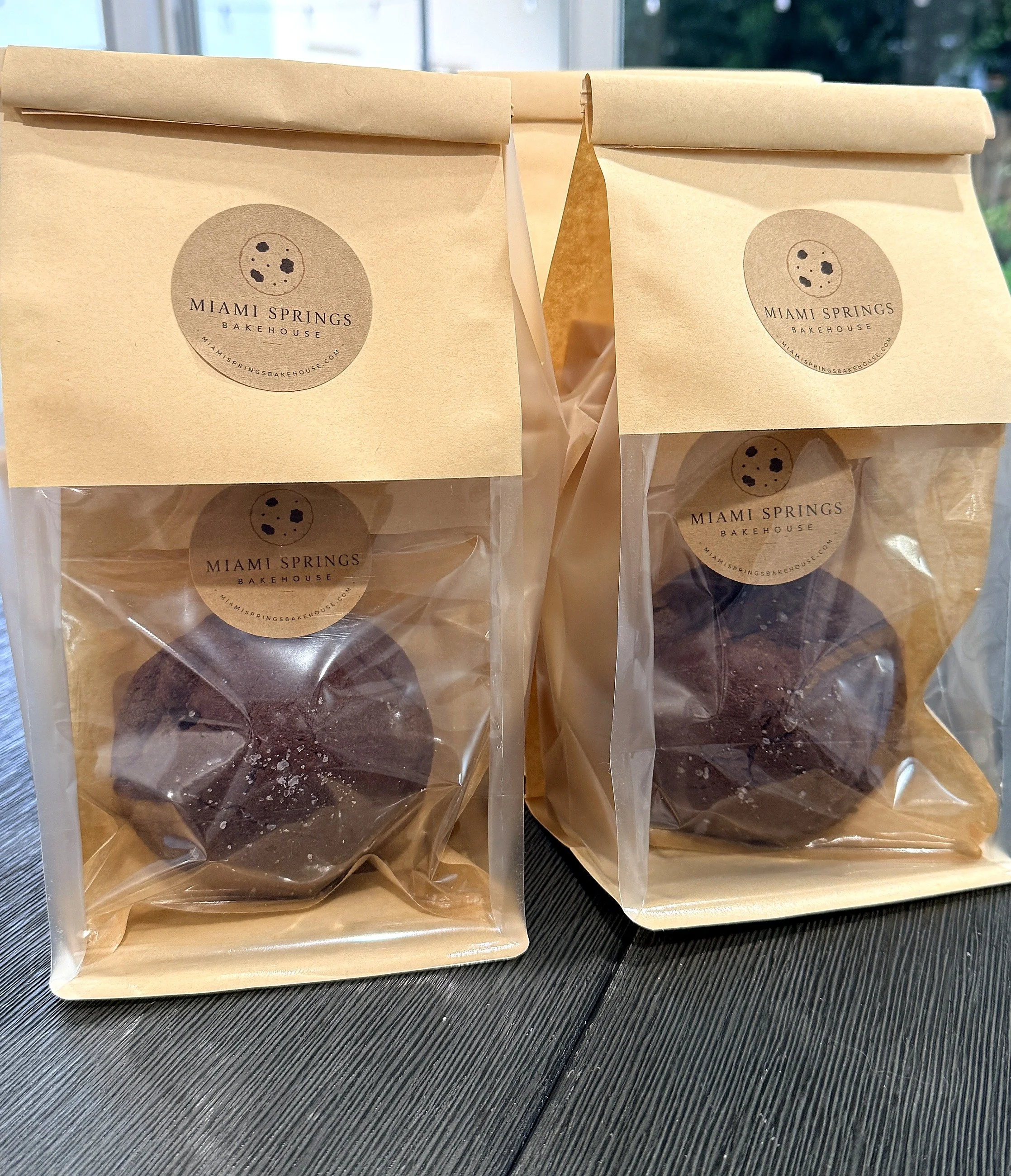 Two paper bags with stickers from Miami Springs Bakehouse, placed on a black textured surface near a window with outdoor scenery in the background.