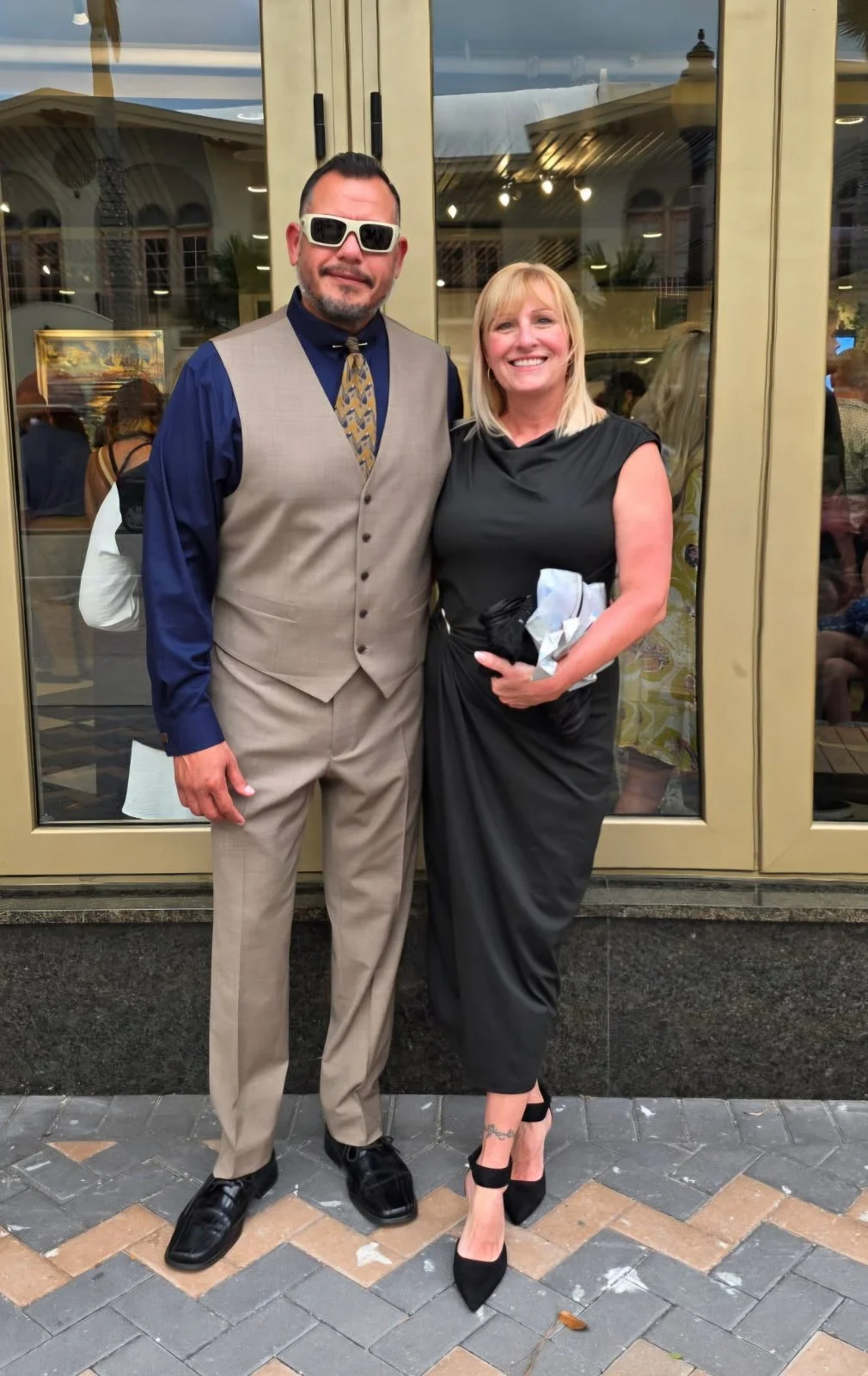 A man and woman standing outside a building, smiling for the camera. The man is wearing a beige vest and pants with a navy shirt and patterned tie, sunglasses, and black shoes. The woman is dressed in a black dress, black heels, and holding a black jacket and some papers, with a tattoo on her ankle.